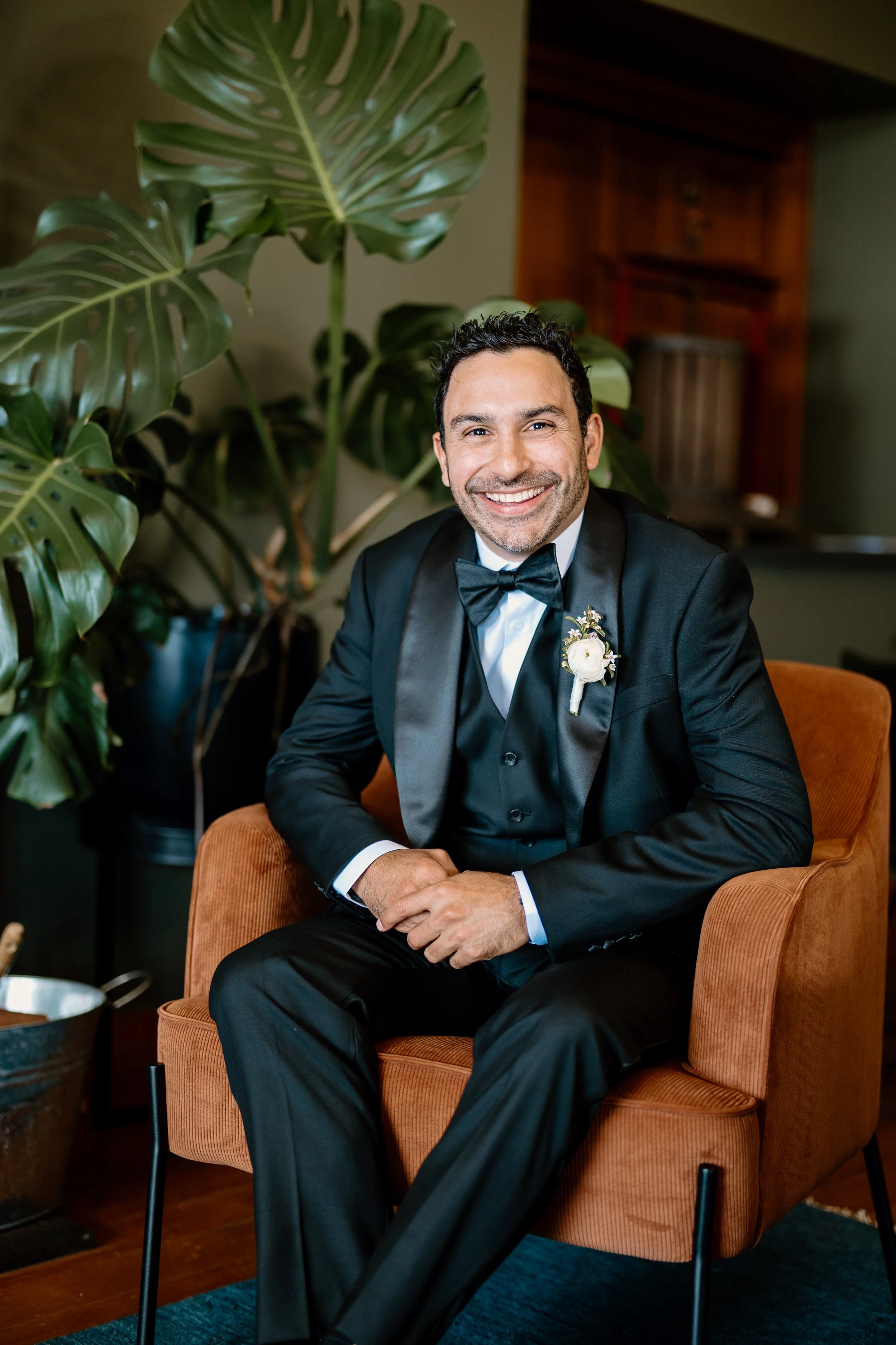A smiling man dressed in a black tuxedo with a bowtie, sitting on an orange armchair. He has a boutonniere on his lapel and is posed in front of a large green leafy plant.