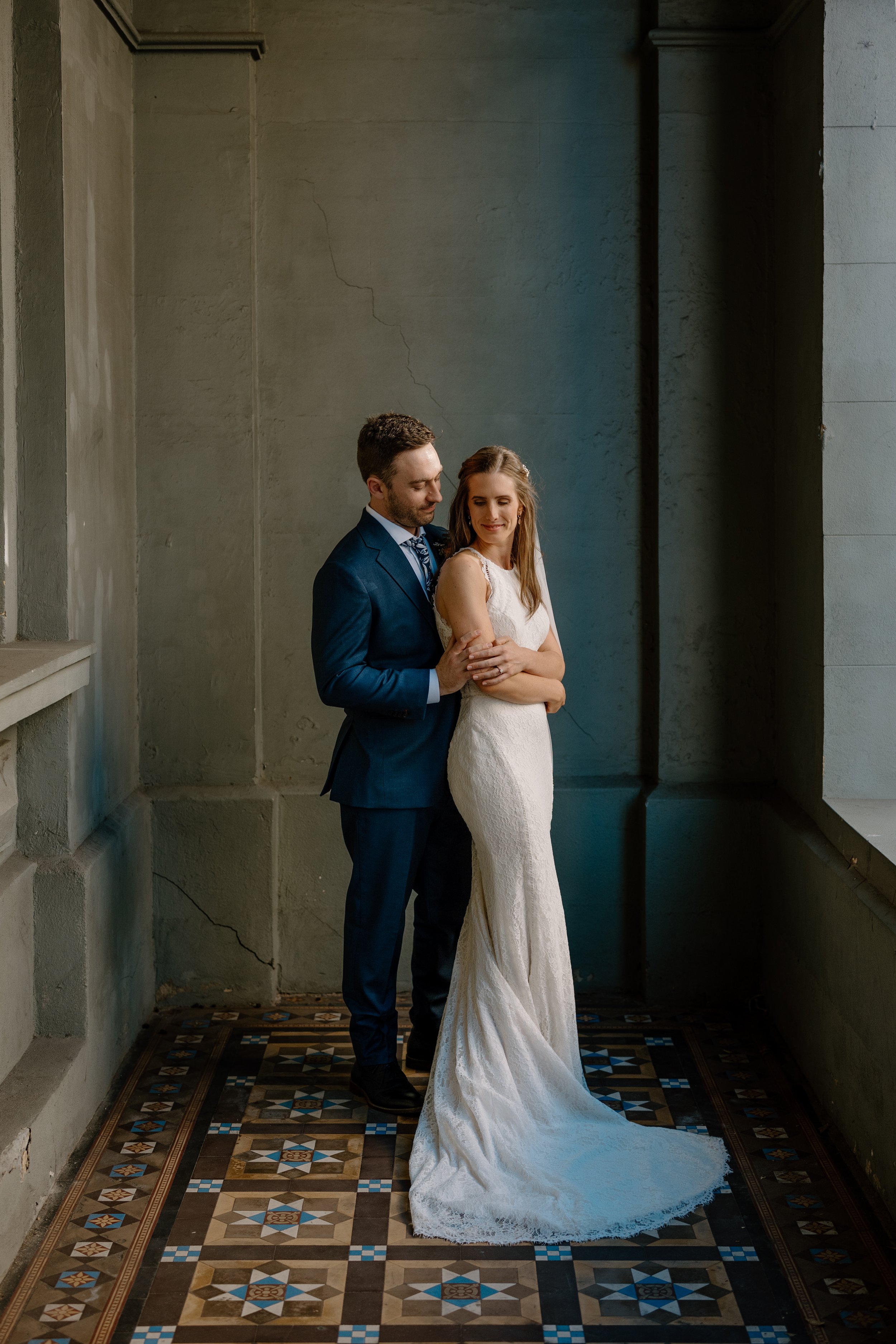 A bride and groom standing together in a dimly lit, industrial-style room with a concrete wall and patterned tile floor in Fremantle