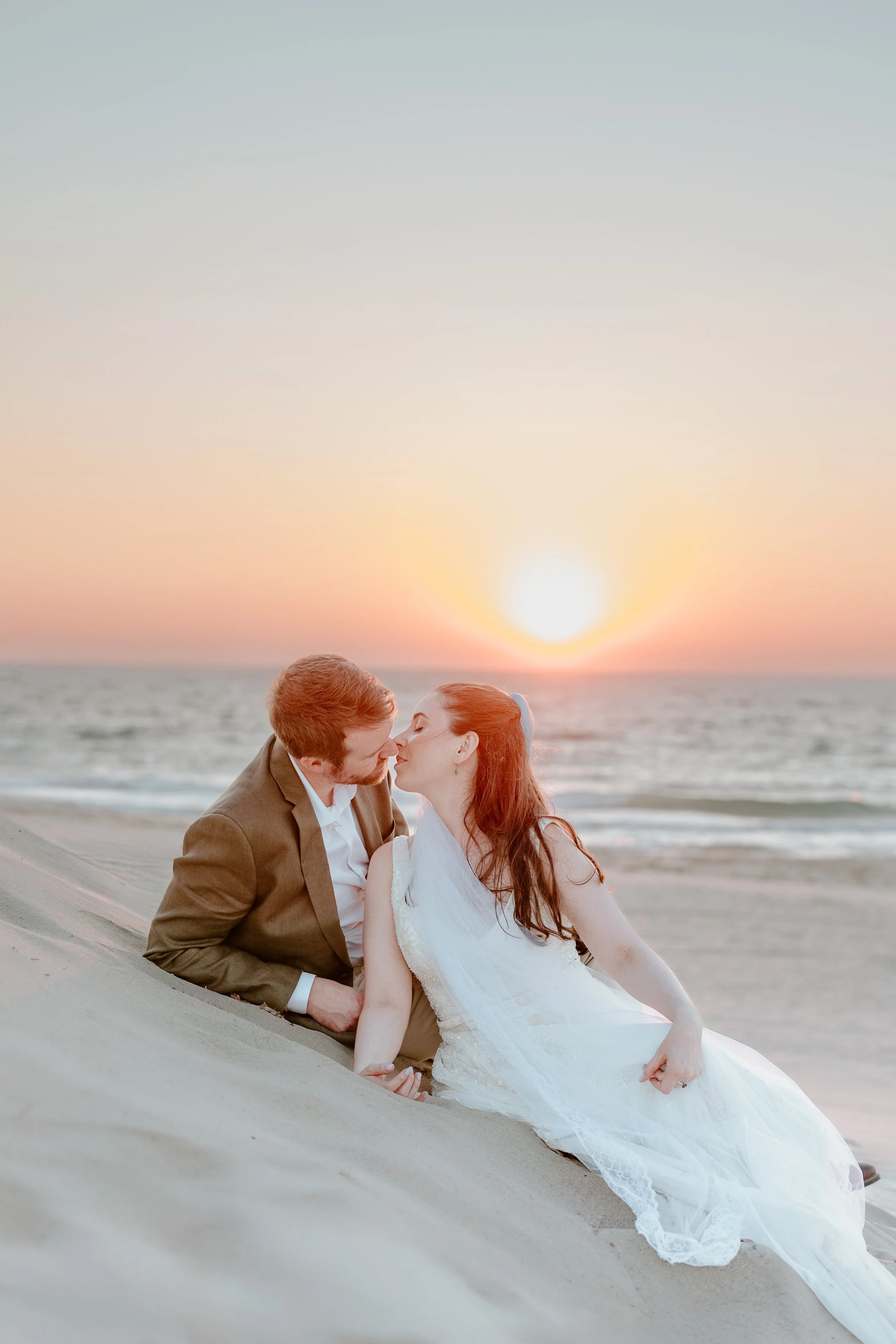A couple in wedding attire sharing a kiss on a beach at sunset.