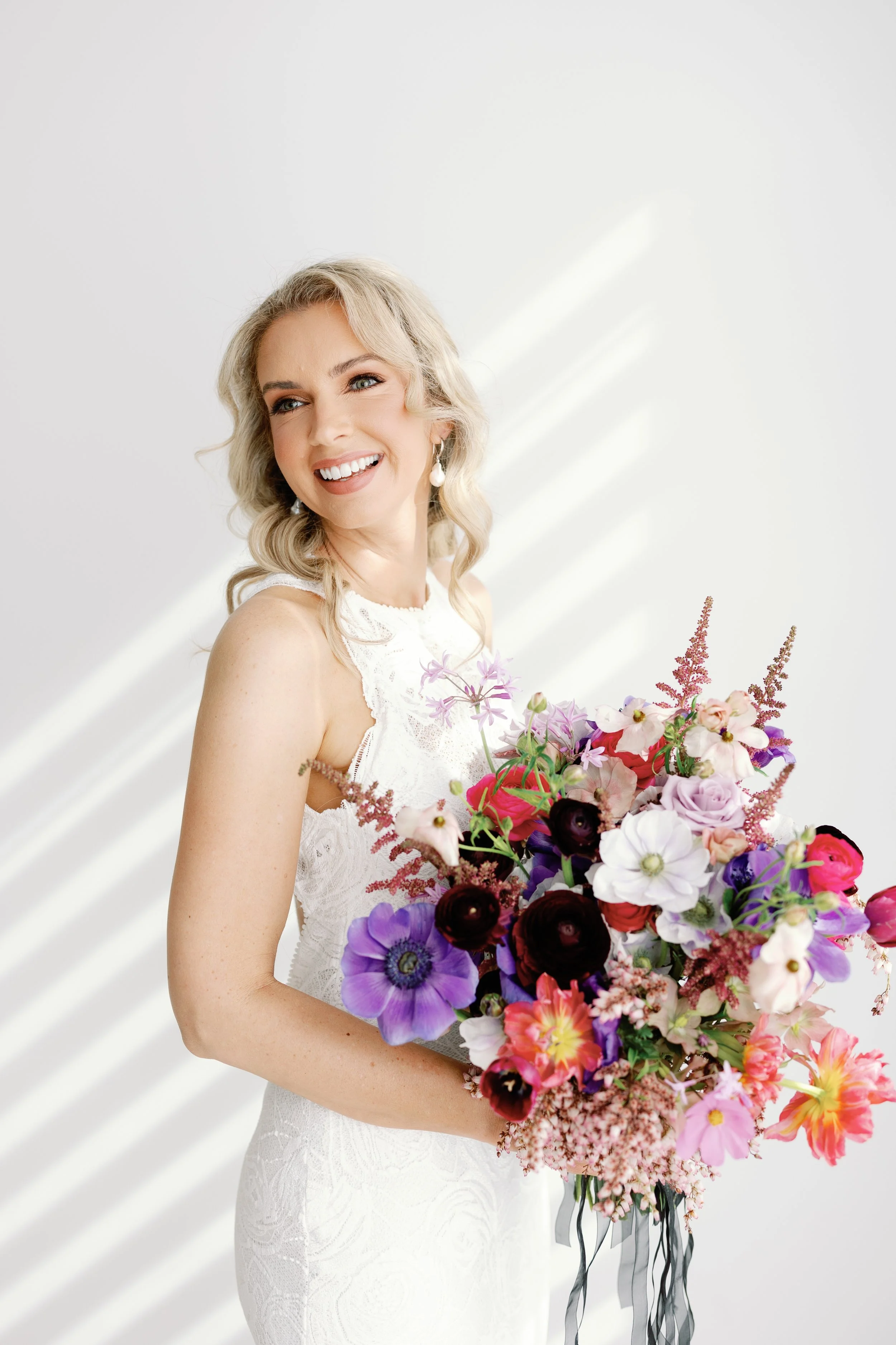 A smiling woman in a white dress holding a large colorful bouquet of flowers with sunlight casting shadows on a white wall.