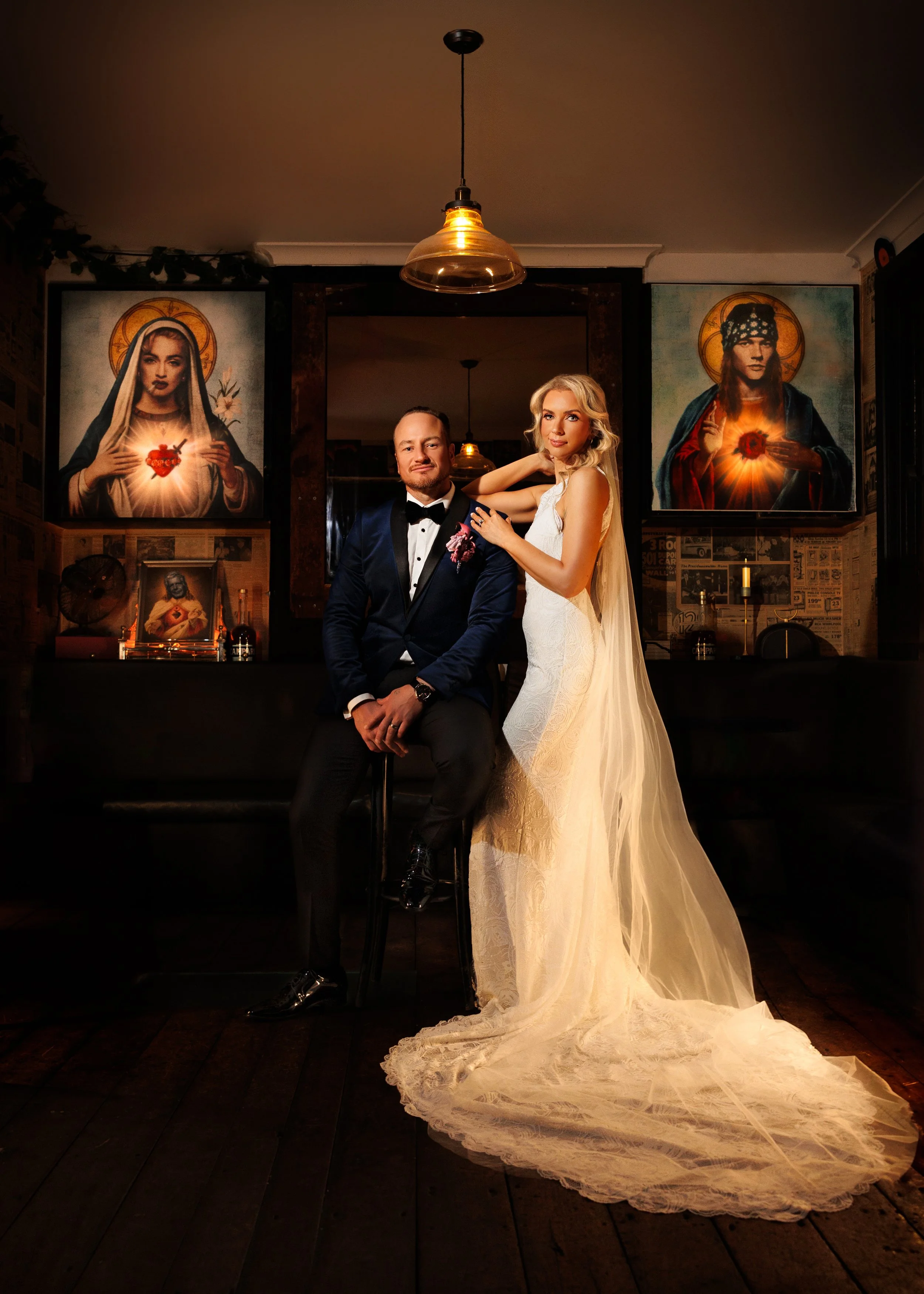 A bride and groom in wedding attire in a dimly lit room with religious artwork of Jesus and Mary in the background.