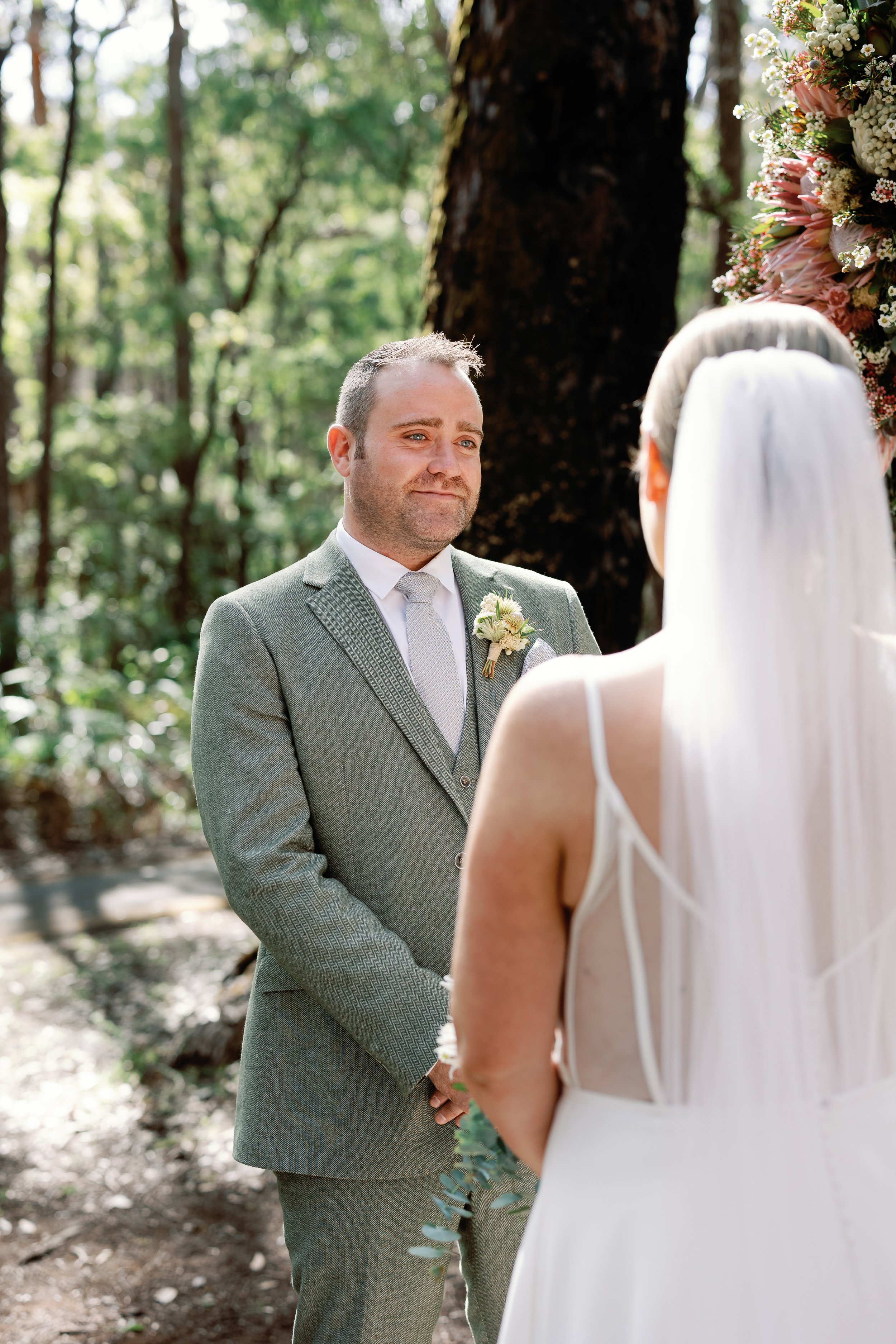 A groom and bride exchanging vows during an outdoor wedding ceremony surrounded by trees, with a floral arrangement visible in the background.