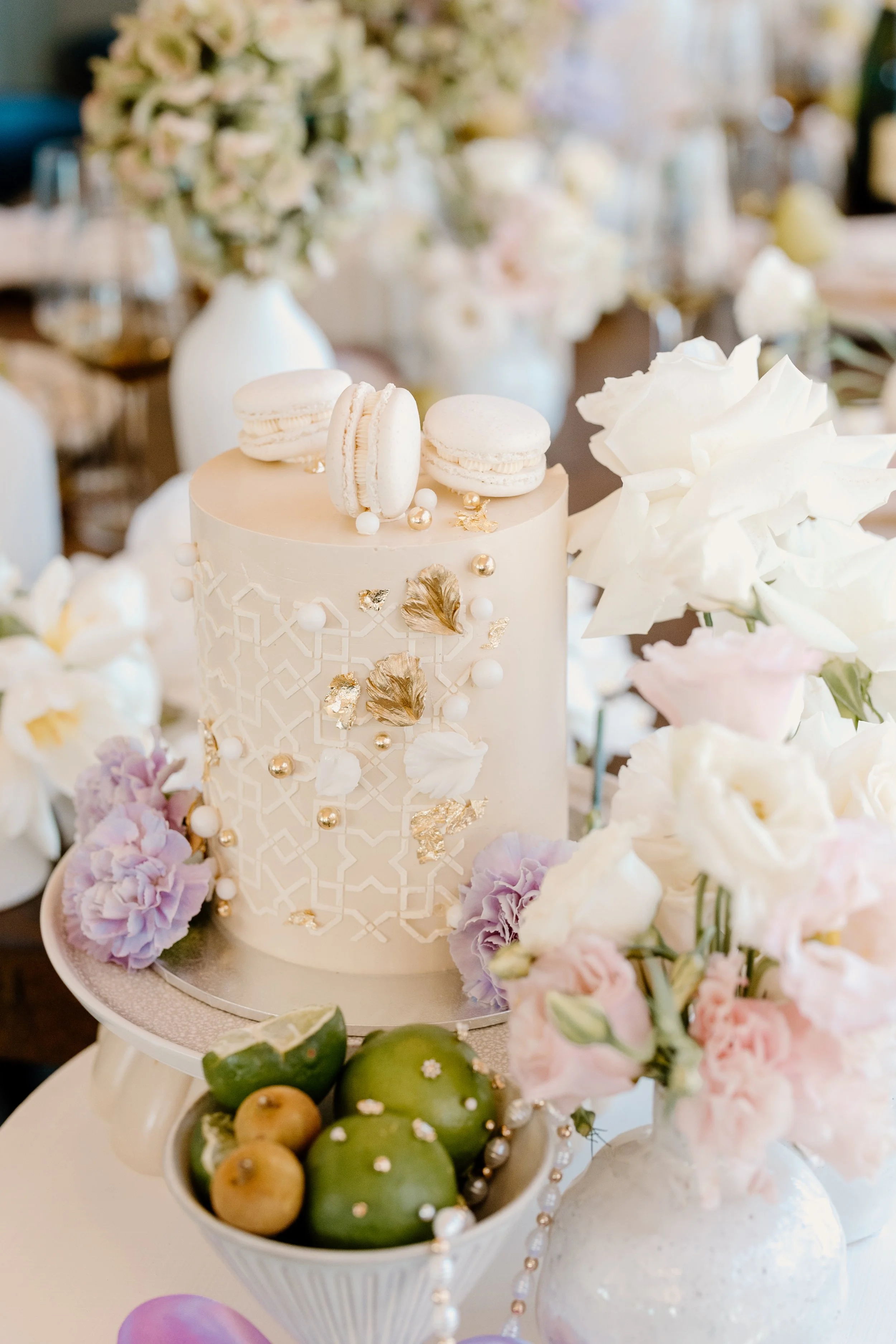A white wedding cake decorated with gold and white accents, topped with three white macarons, surrounded by floral arrangements in shades of white, pink, and purple, with green fruits at the base.