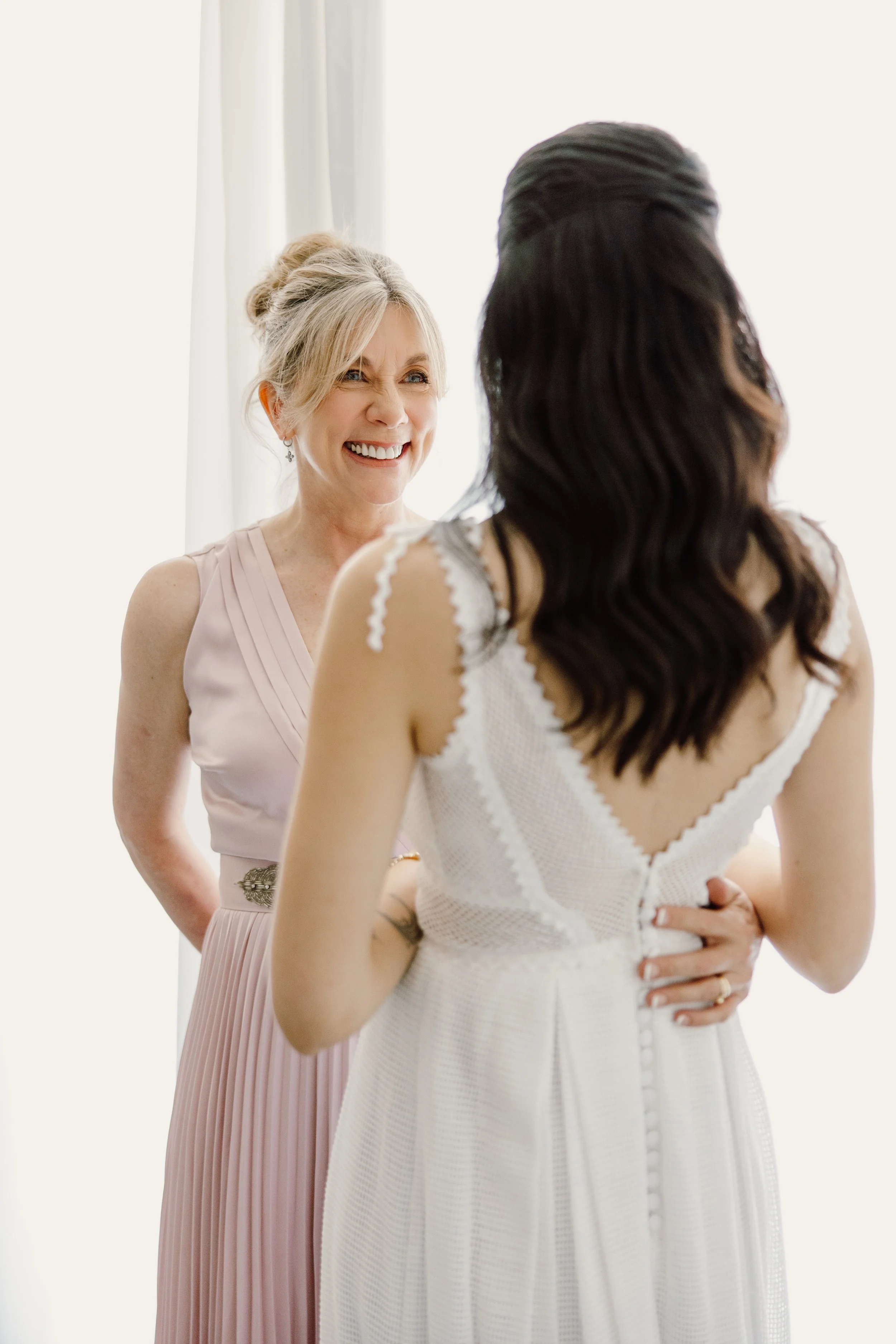 A bride in a white dress with buttons on the back, and a woman in a light pink dress smiling and talking to her.