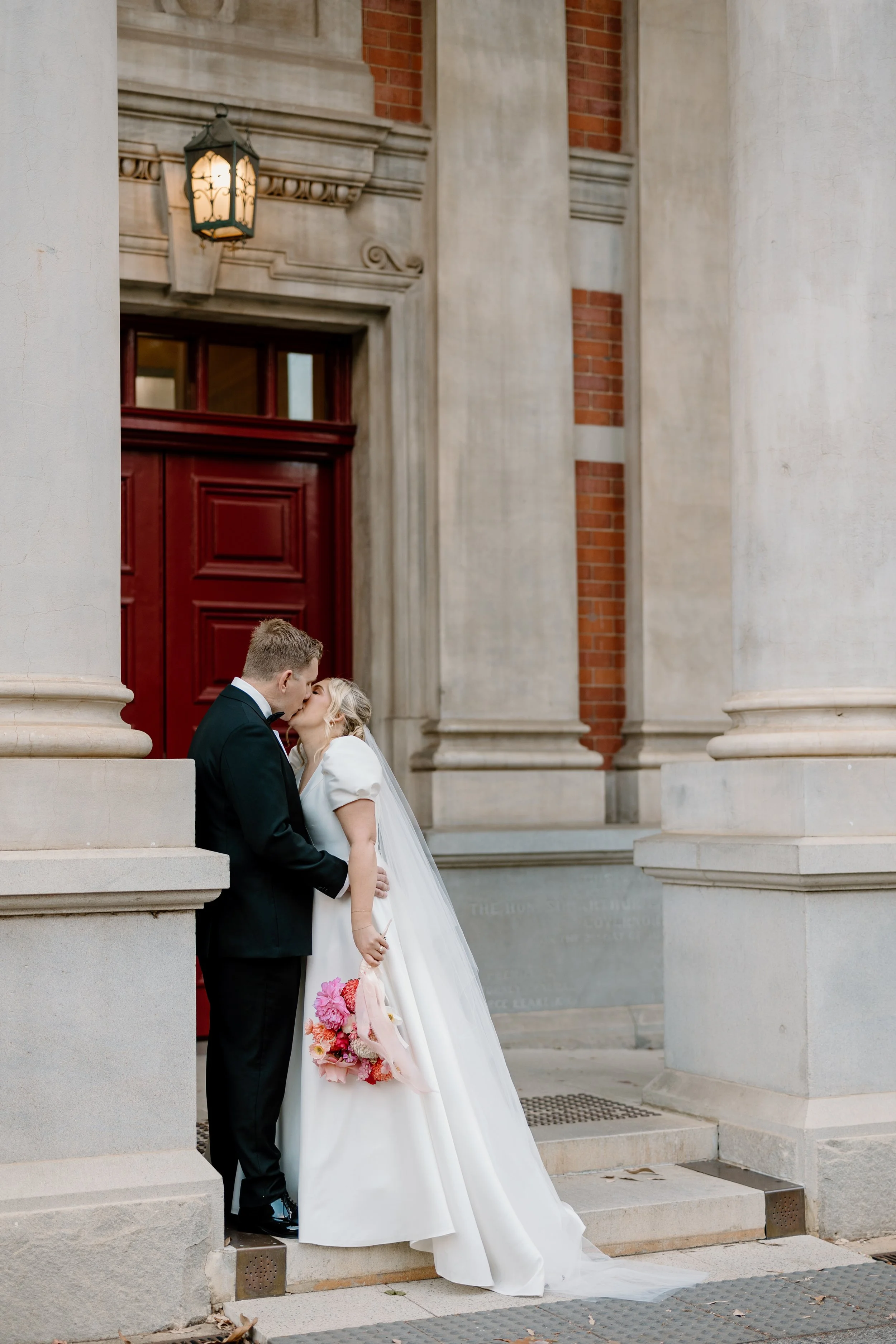 A bride and groom kissing outside a building with large stone columns and a red door, the bride holding a pink and red bouquet at Como Treasury Perth 