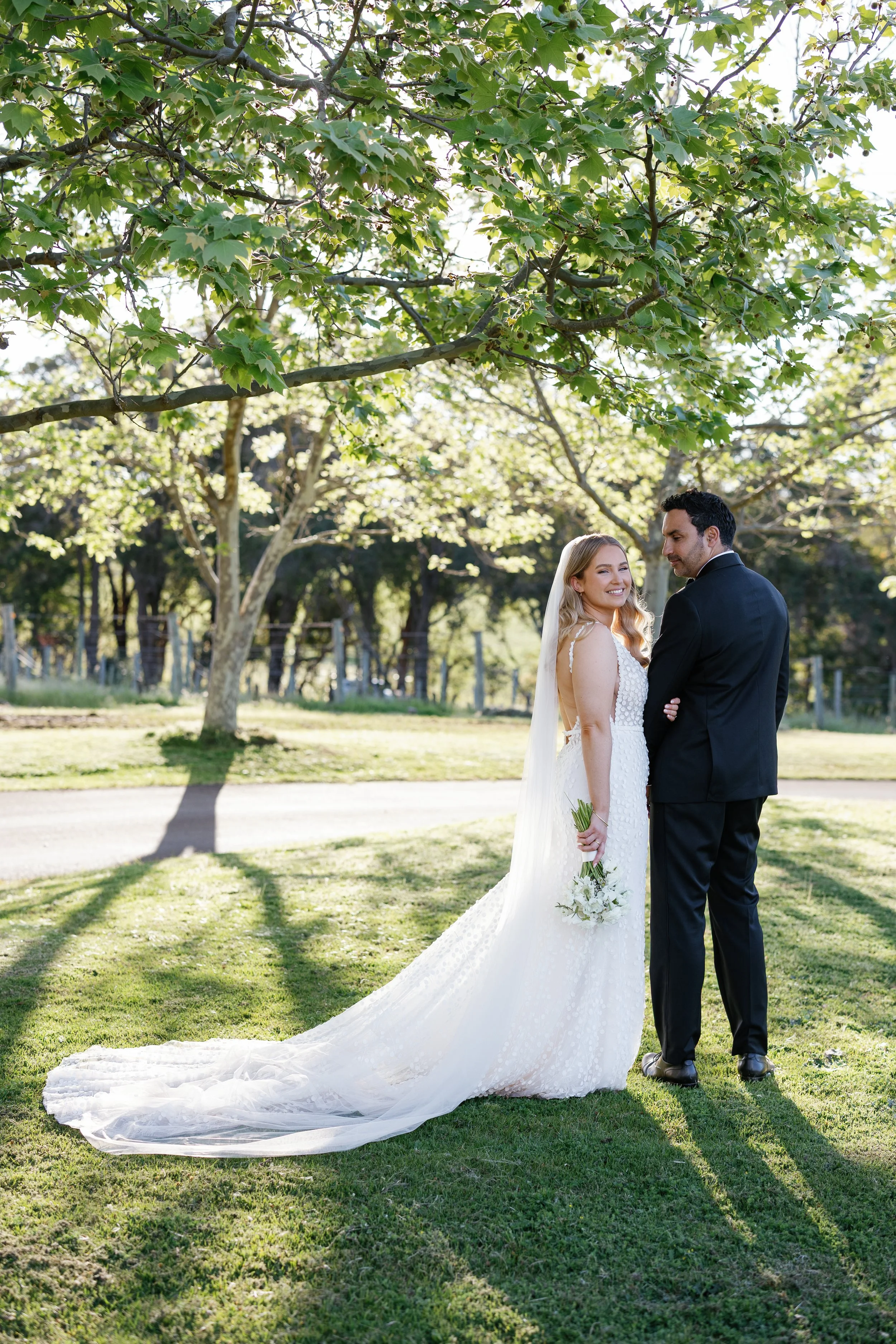 A bride and groom standing together under a tree in a park, with sunlight filtering through the leaves. The bride is smiling and holding a bouquet of white flowers, while the groom looks at her affectionately. The bride is dressed in a white wedding 