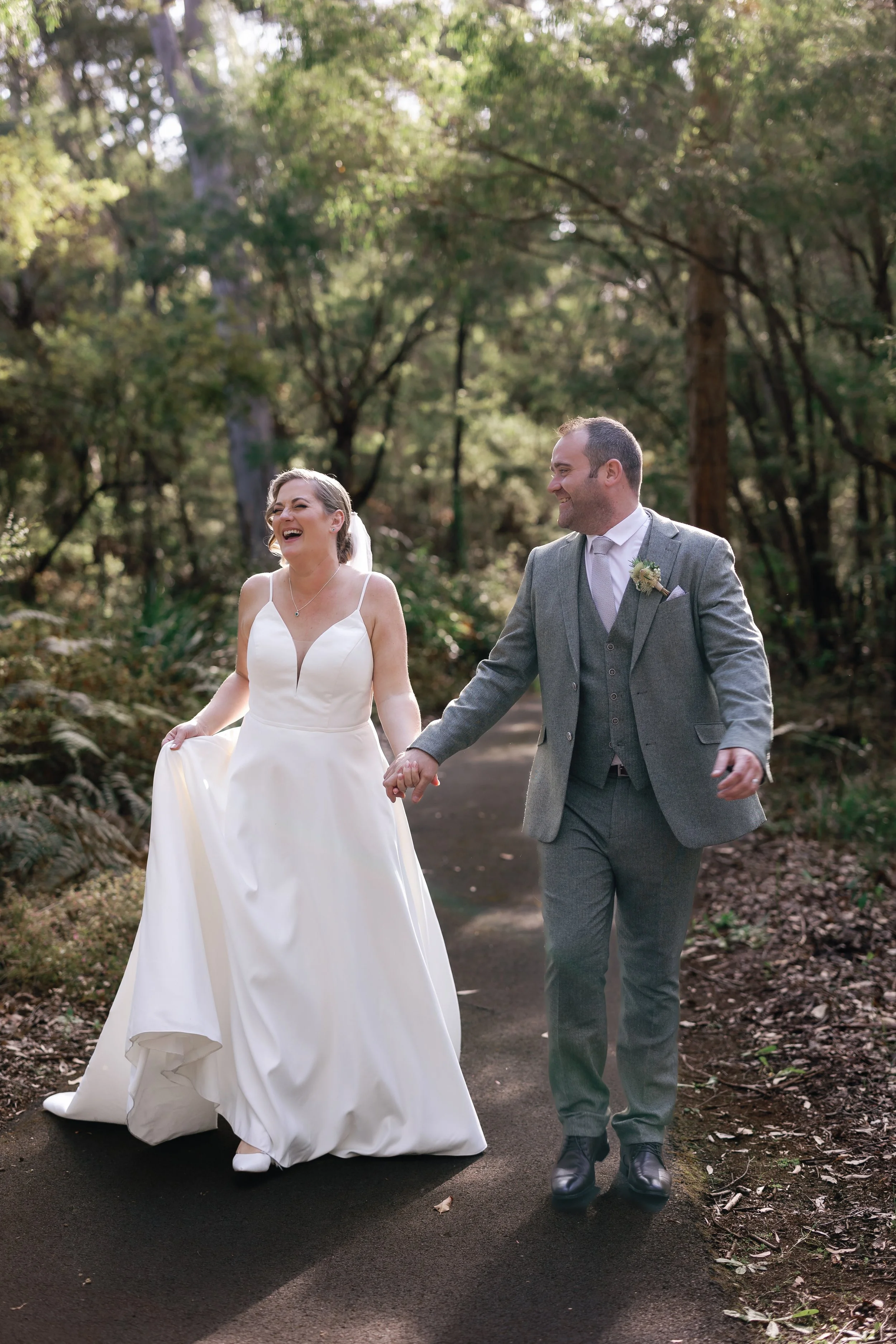 A bride and groom walking hand in hand on a wooded path, smiling and laughing.