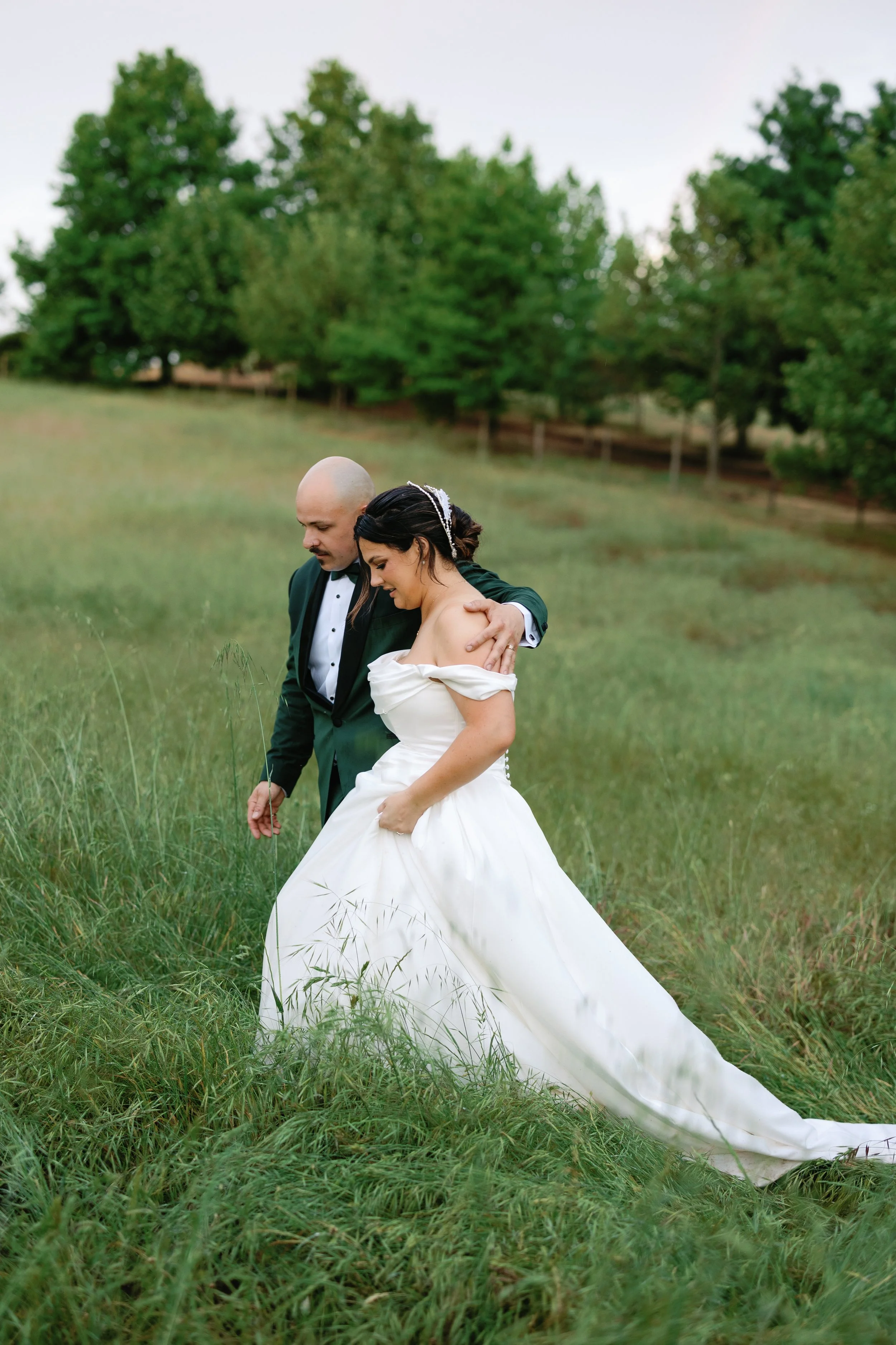 A bride and groom walking together on a grassy field, with the groom gently holding the bride's shoulders, surrounded by green trees in Margaret River