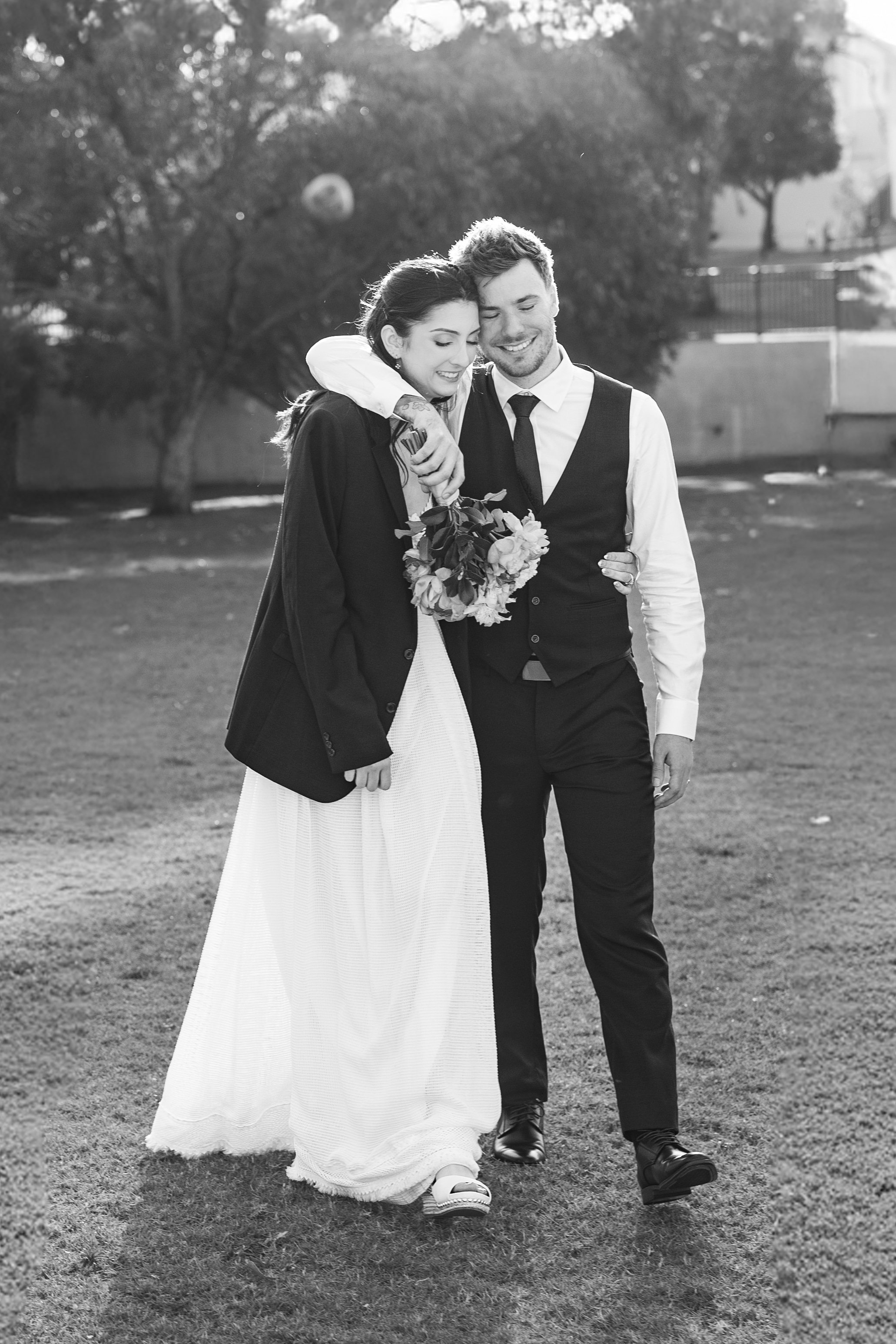 Black and white photo of a smiling couple walking outdoors, the woman holding a bouquet of flowers and wearing a long dress and blazer, while the man wears a vest, shirt, and tie.