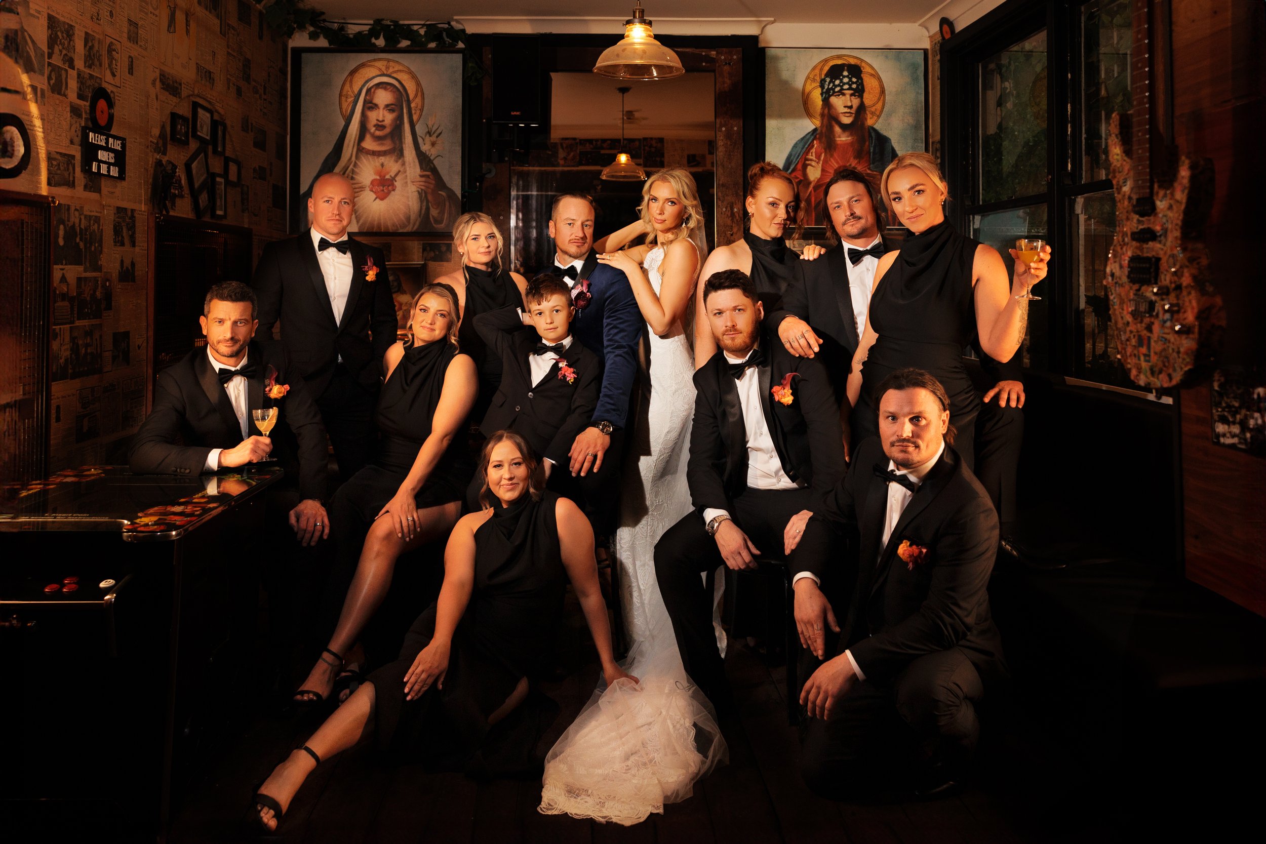 Group of people dressed in formal attire, likely at a wedding reception, in a dimly lit restaurant or bar with religious artwork on the walls.