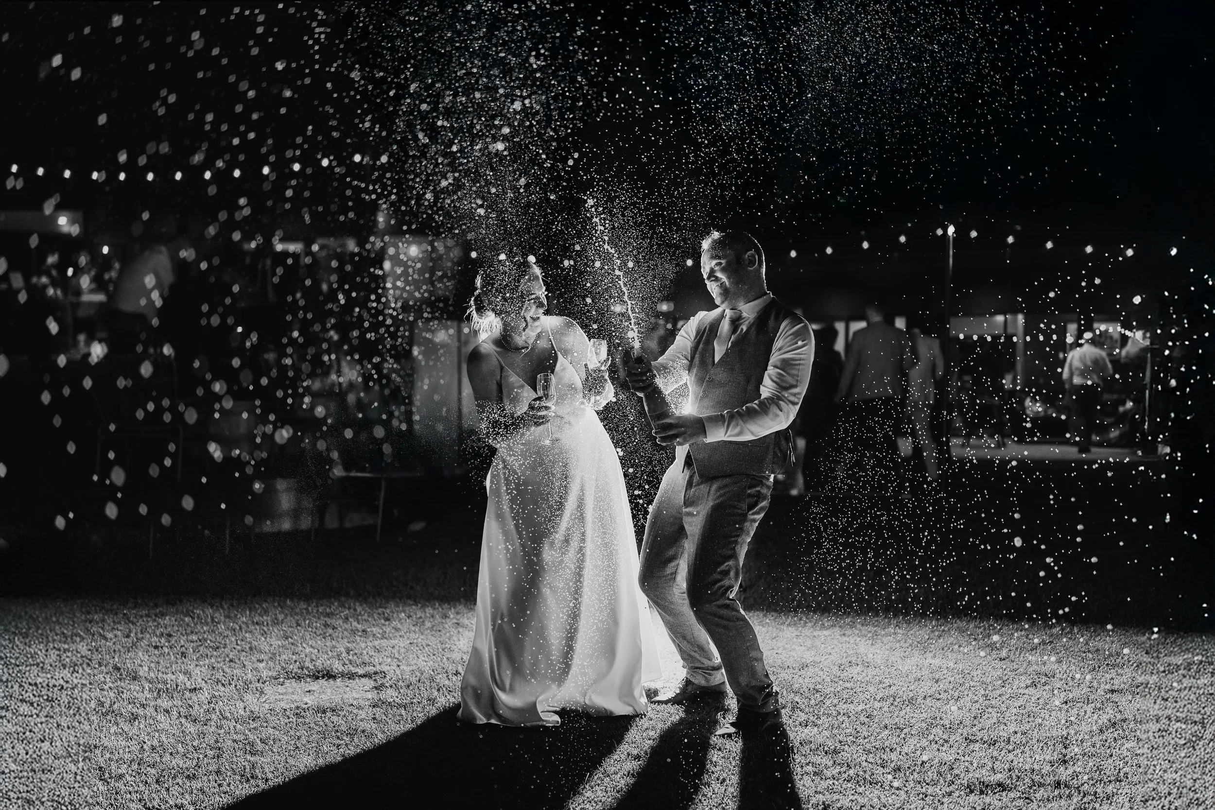A black-and-white photo of a bride and groom dancing at their wedding reception, holding champagne glasses, while being sprayed with champagne at Tanah Marah Margaret River