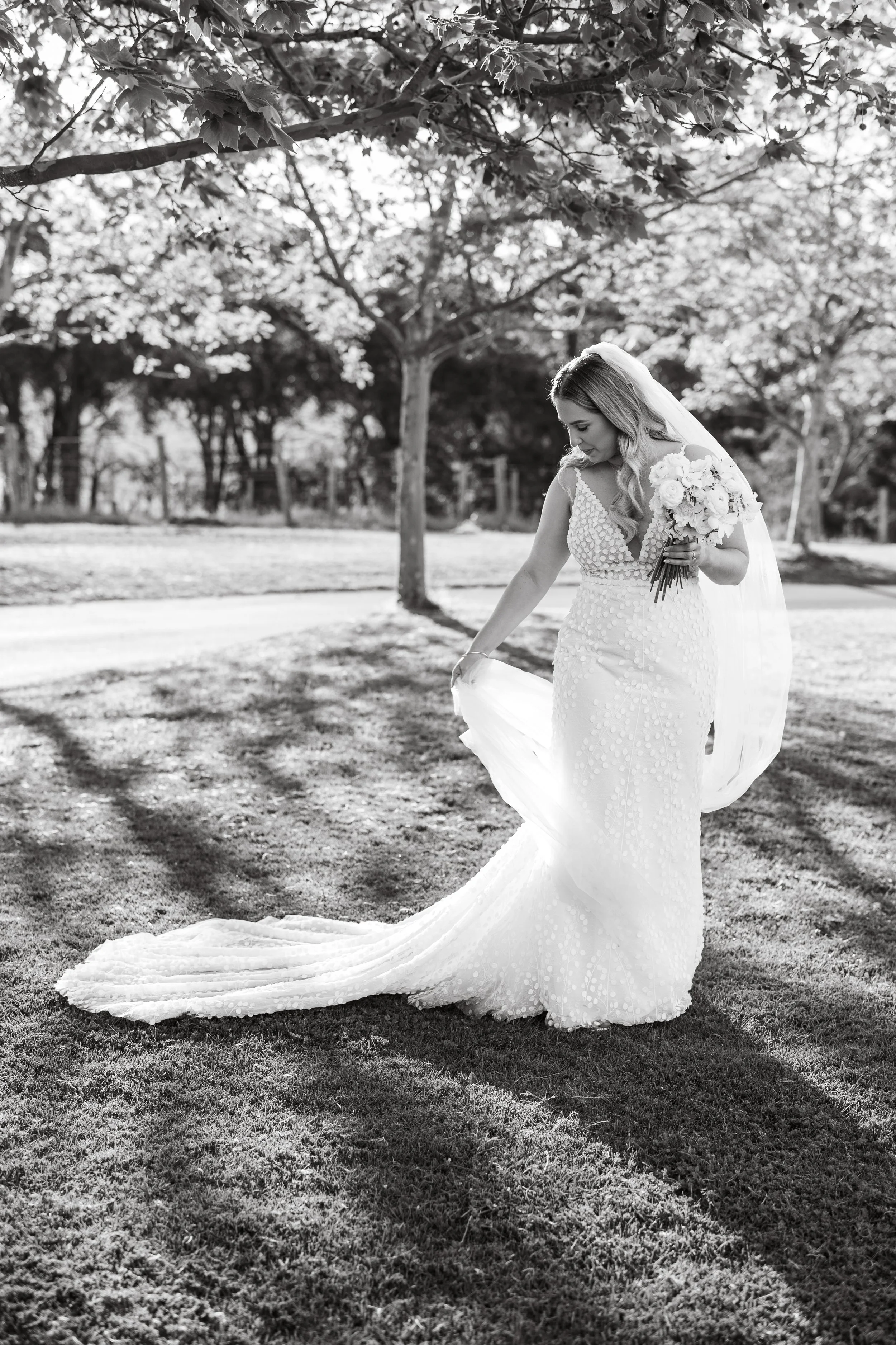 A bride in a white wedding dress holding a bouquet of flowers, standing outdoors on grass with trees in the background, in black and white.
