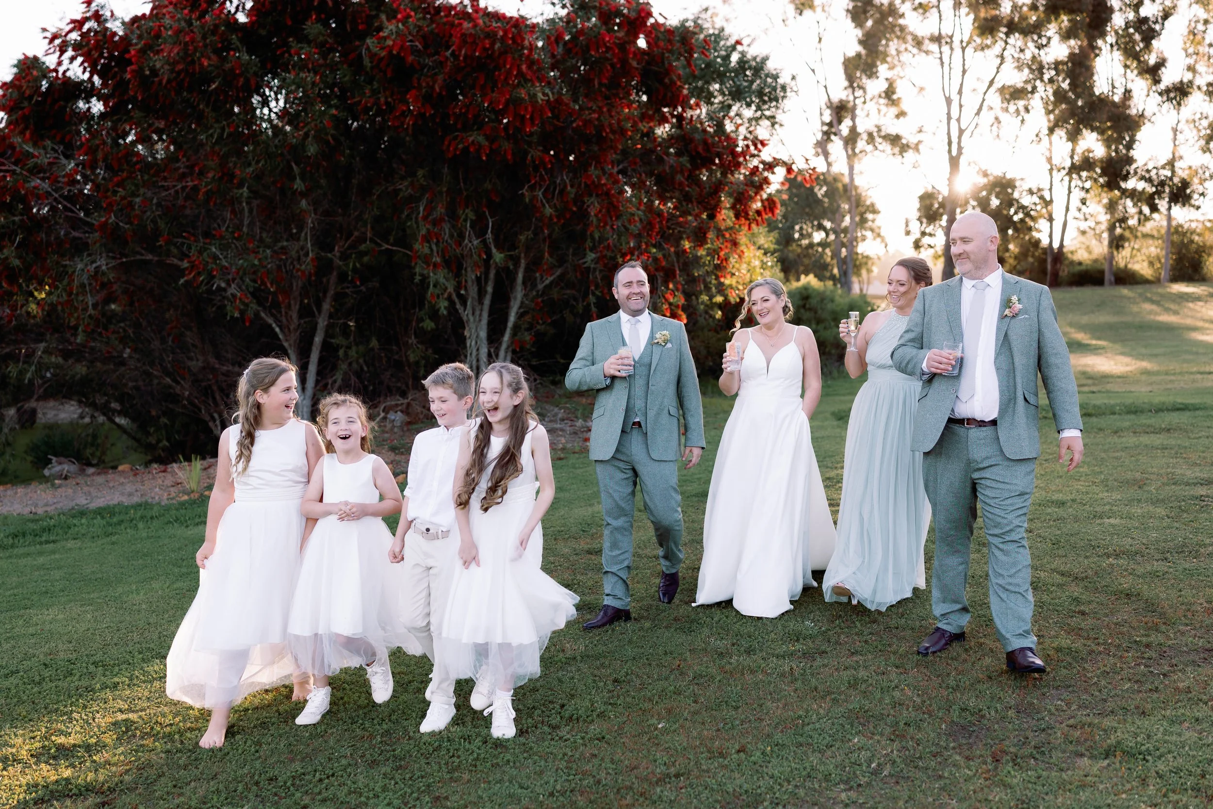 A wedding group walking outdoors on a grassy area at sunset, including the bride in a white wedding dress, the groom and two bridesmaids in suits, and four young girls in white dresses, all smiling and holding glasses of champagne or wine.