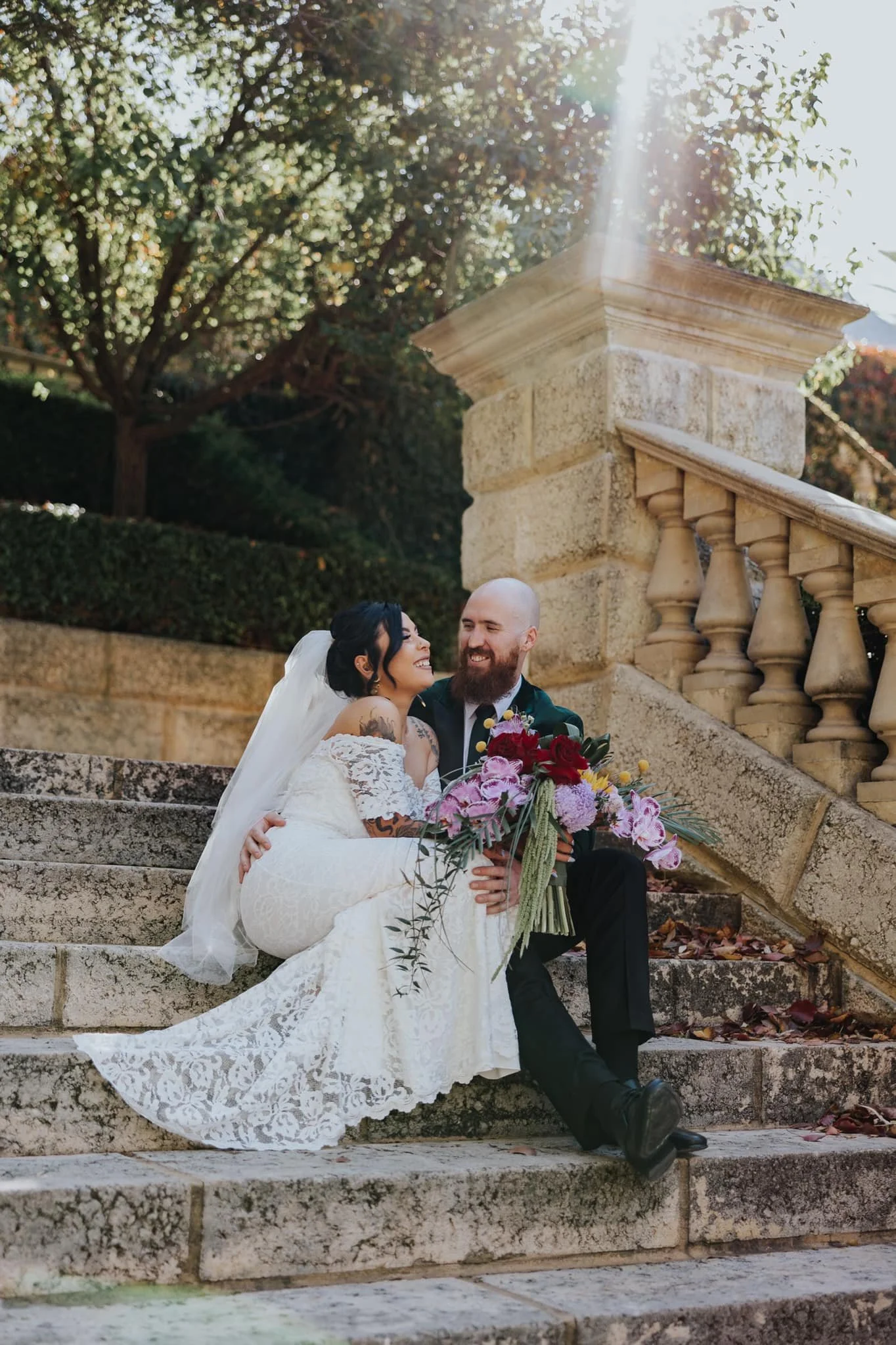 A newlywed couple sitting on stone steps outdoors. The bride is in a white lace wedding dress with a veil, holding a vibrant bouquet. The groom is in a dark suit with a boutonniere. Both are smiling and looking at each other, with sunlight filtering 
