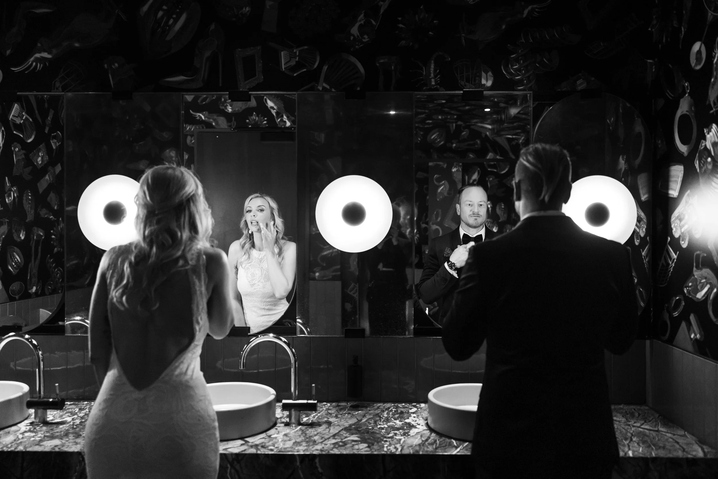 A woman in a wedding dress and a man in a tuxedo stand in front of mirrors in a bathroom, getting ready, with bright round lights illuminating the space.