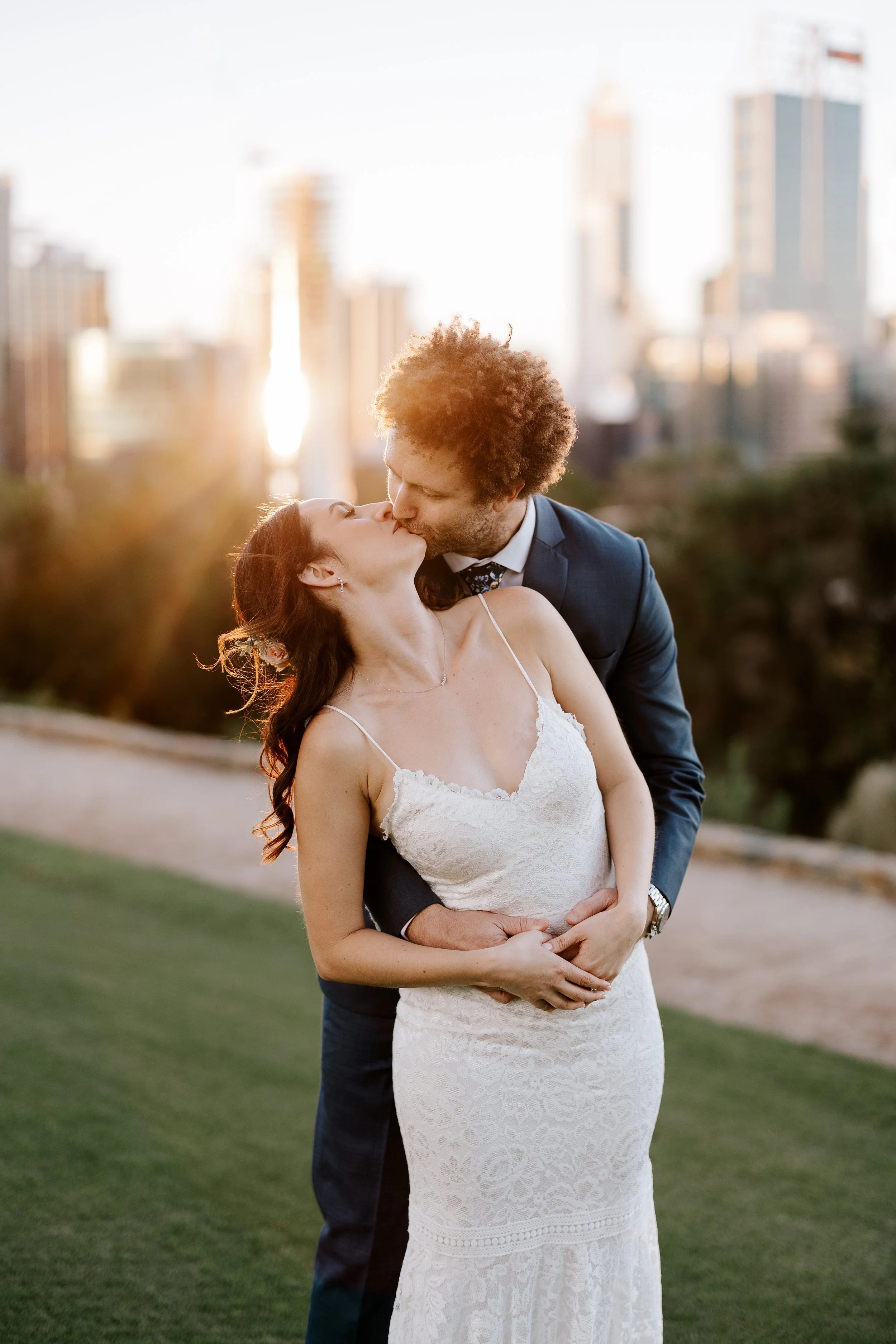 A newlywed couple sharing a kiss outdoors at sunset, with city skyline in the background.
