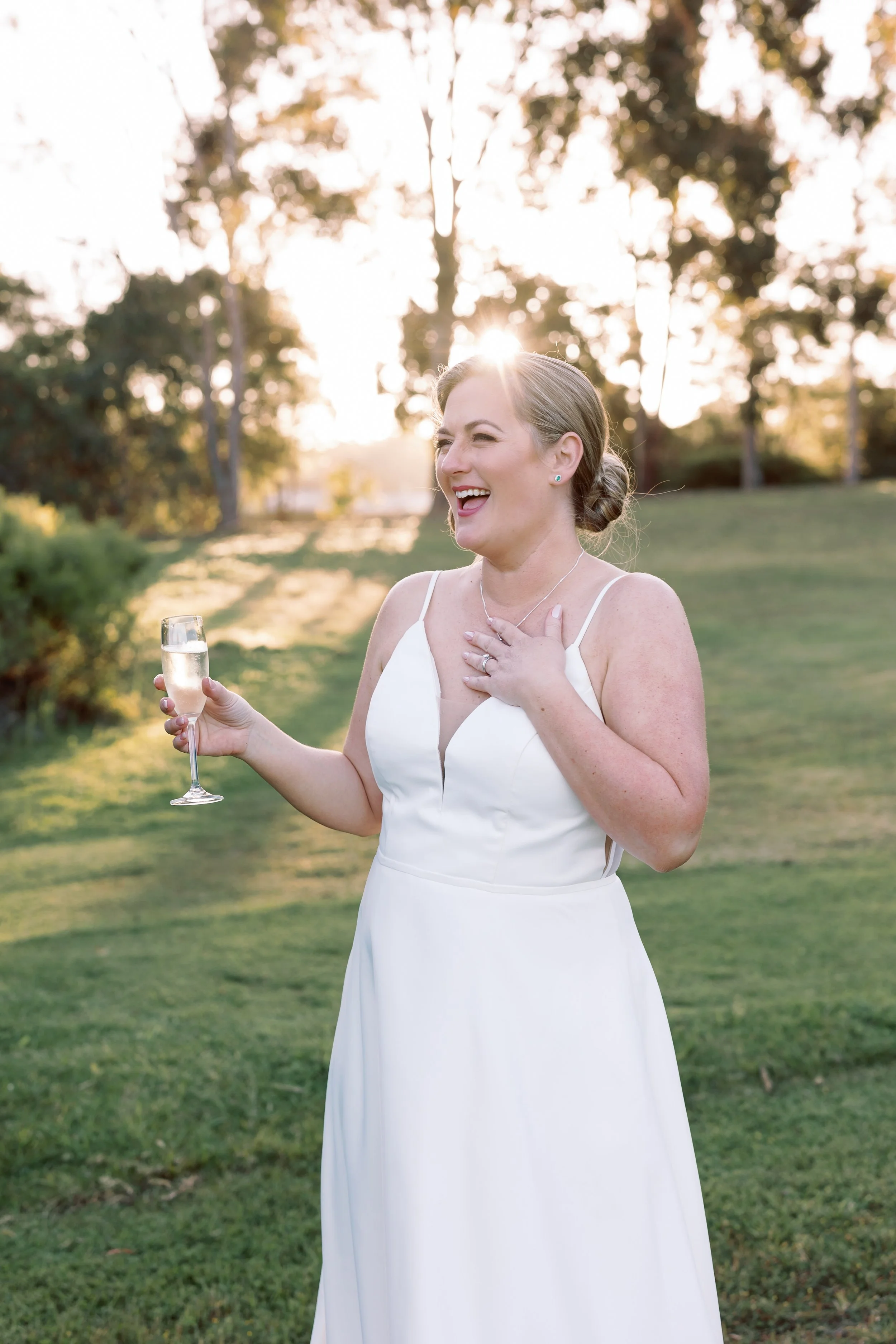 A bride in a white wedding dress holding a glass of champagne, smiling and touching her chest, standing outdoors in a grassy area with trees and the setting sun in the background.