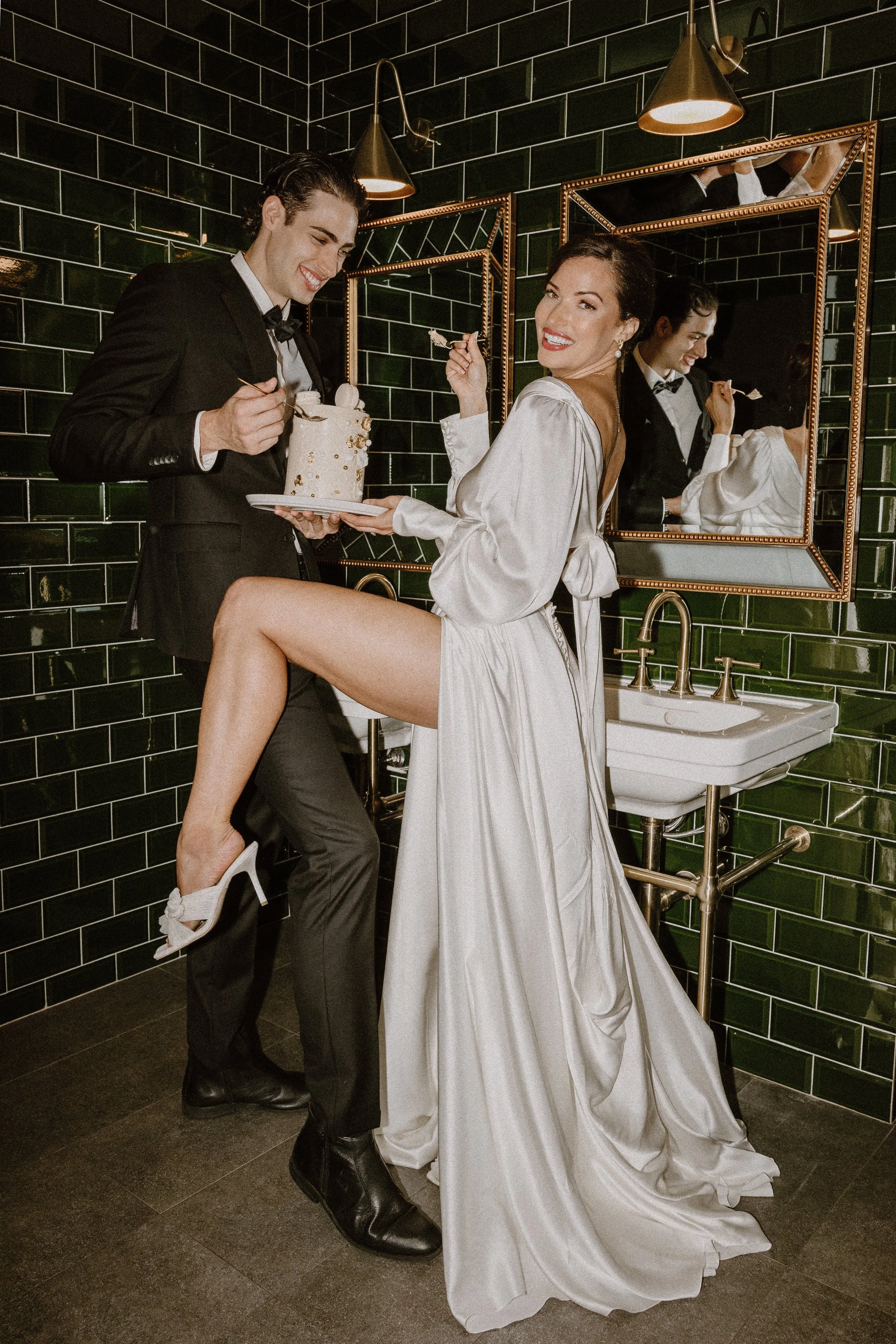 A woman in a long white satin dress with puffed sleeves and a man in a black tuxedo with a bow tie in a bathroom with dark green tiled walls. The woman has one leg raised and is smiling at the camera while holding a fork. The man is holding a plate with a small cake and a fork. The bathroom has a white sink, gold lighting fixtures, and mirrors reflecting the scene.