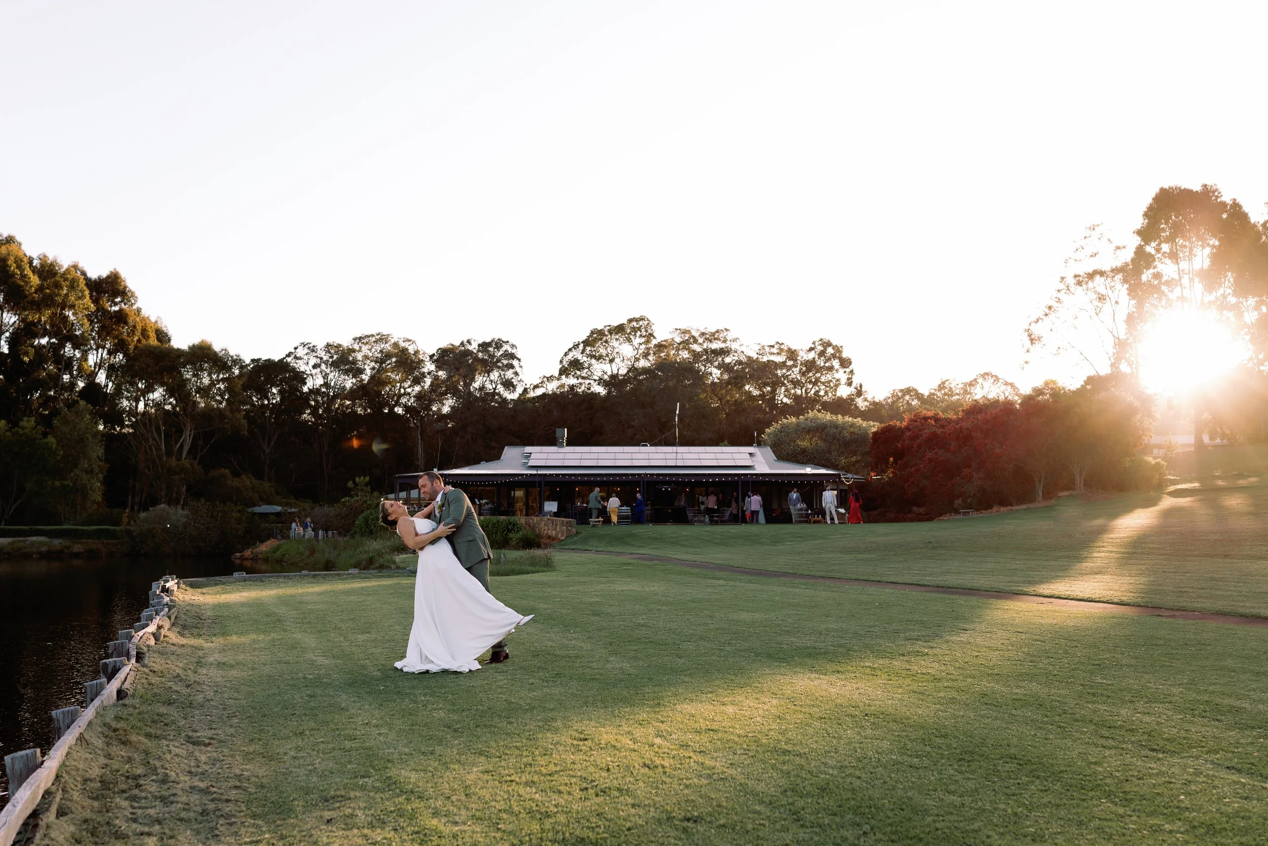 A bride and groom dancing on a grassy area near a small lake during sunset at a wedding celebration, with a large building and trees in the background.