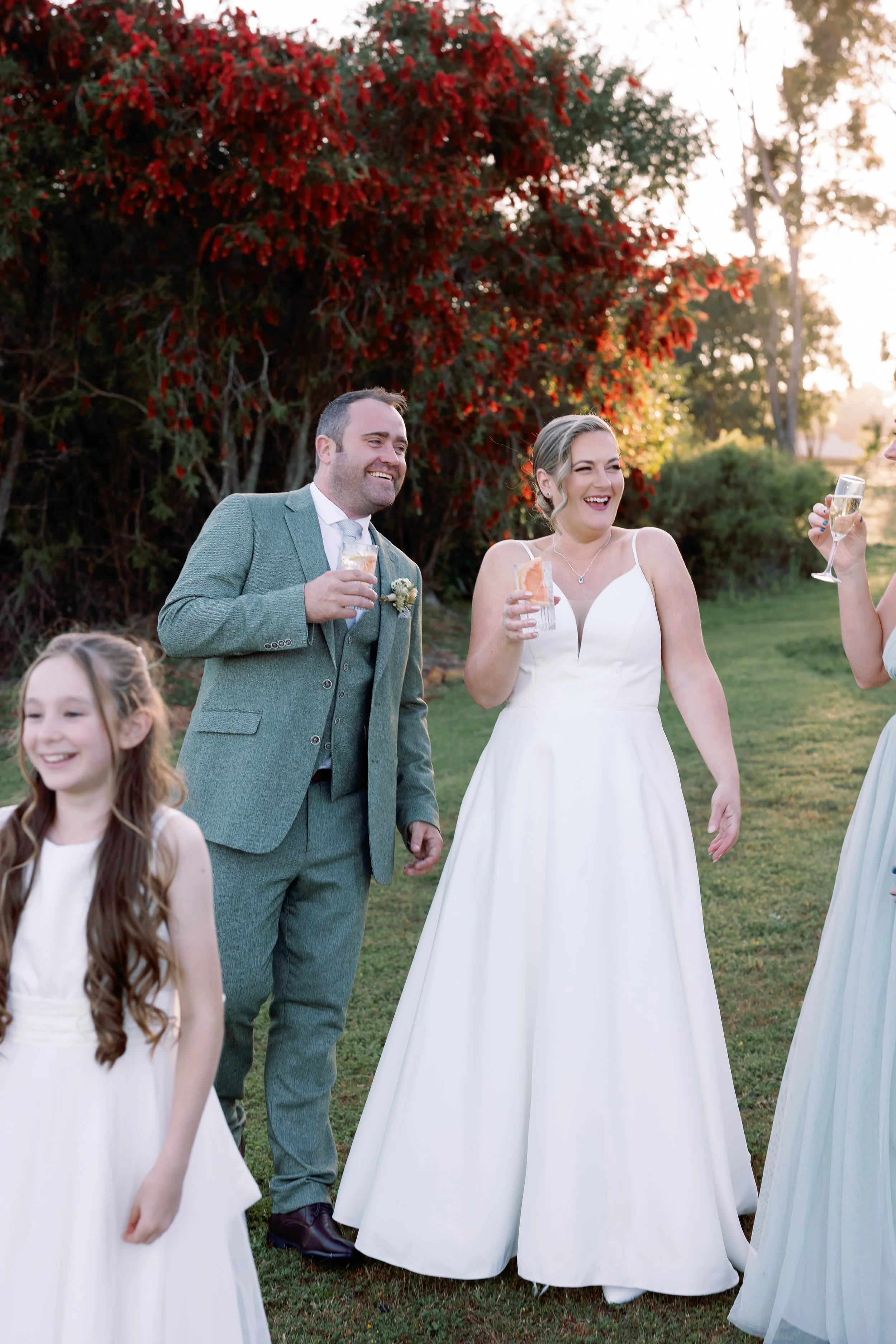 A wedding celebration outdoors with a bride, groom, and two women and a girl, all holding glasses of champagne and smiling, with trees and the setting sun in the background.