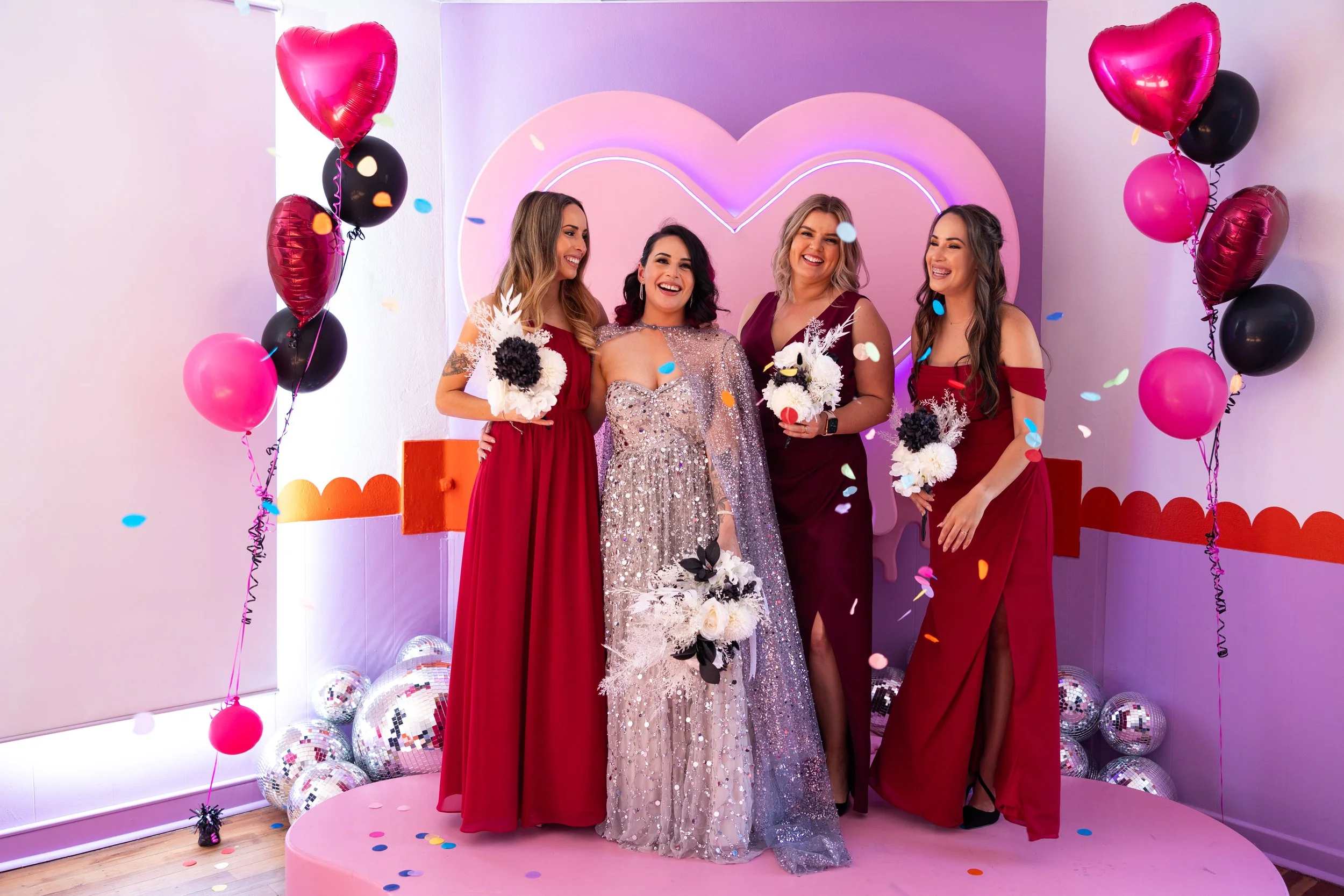 Four women in red dresses and one woman in a silver, sequined gown standing on a pink platform with balloons, confetti, and a pink heart-shaped neon sign in the background, at a celebration or party.