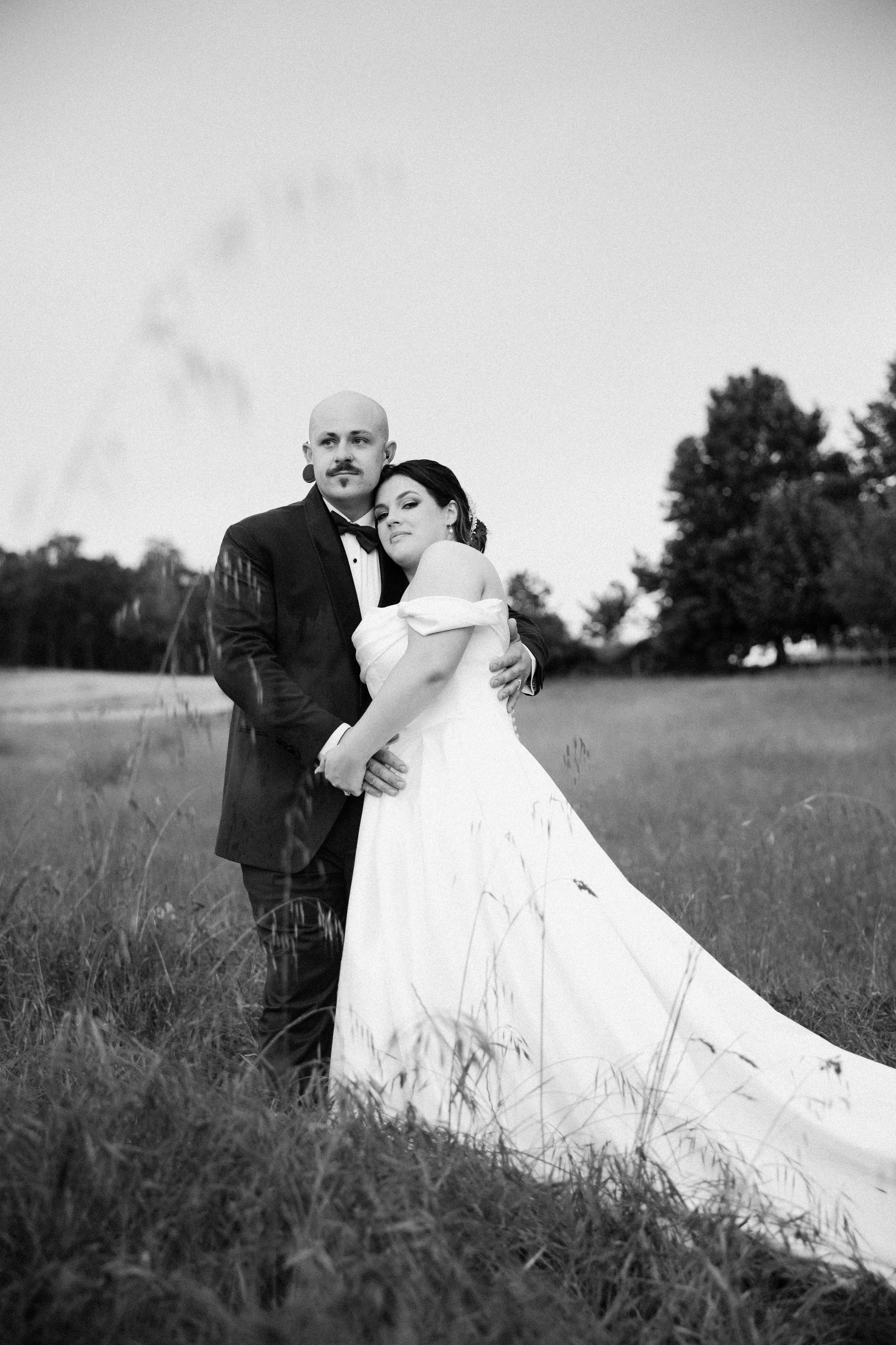 Black and white photo of a bride and groom standing in a field. The groom is wearing a tuxedo with a bow tie and the bride is in a strapless wedding dress. They are embracing each other, with a few trees in the background.