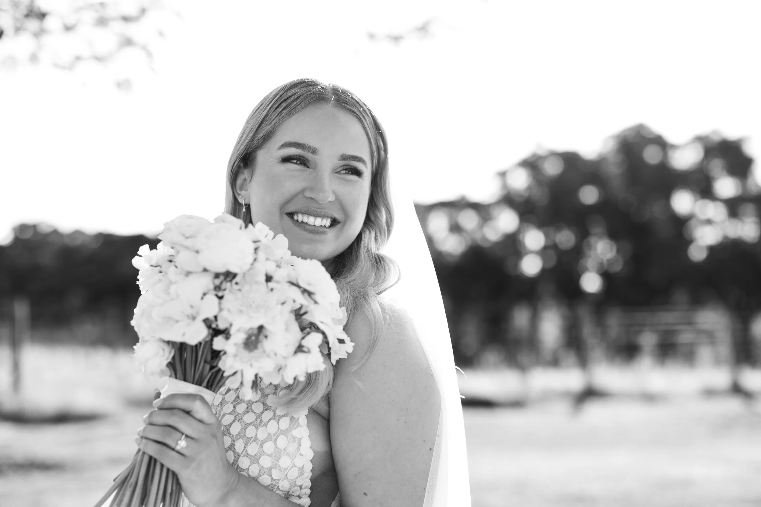 Black and white photo of a smiling bride holding a bouquet of flowers outdoors with trees in the background.