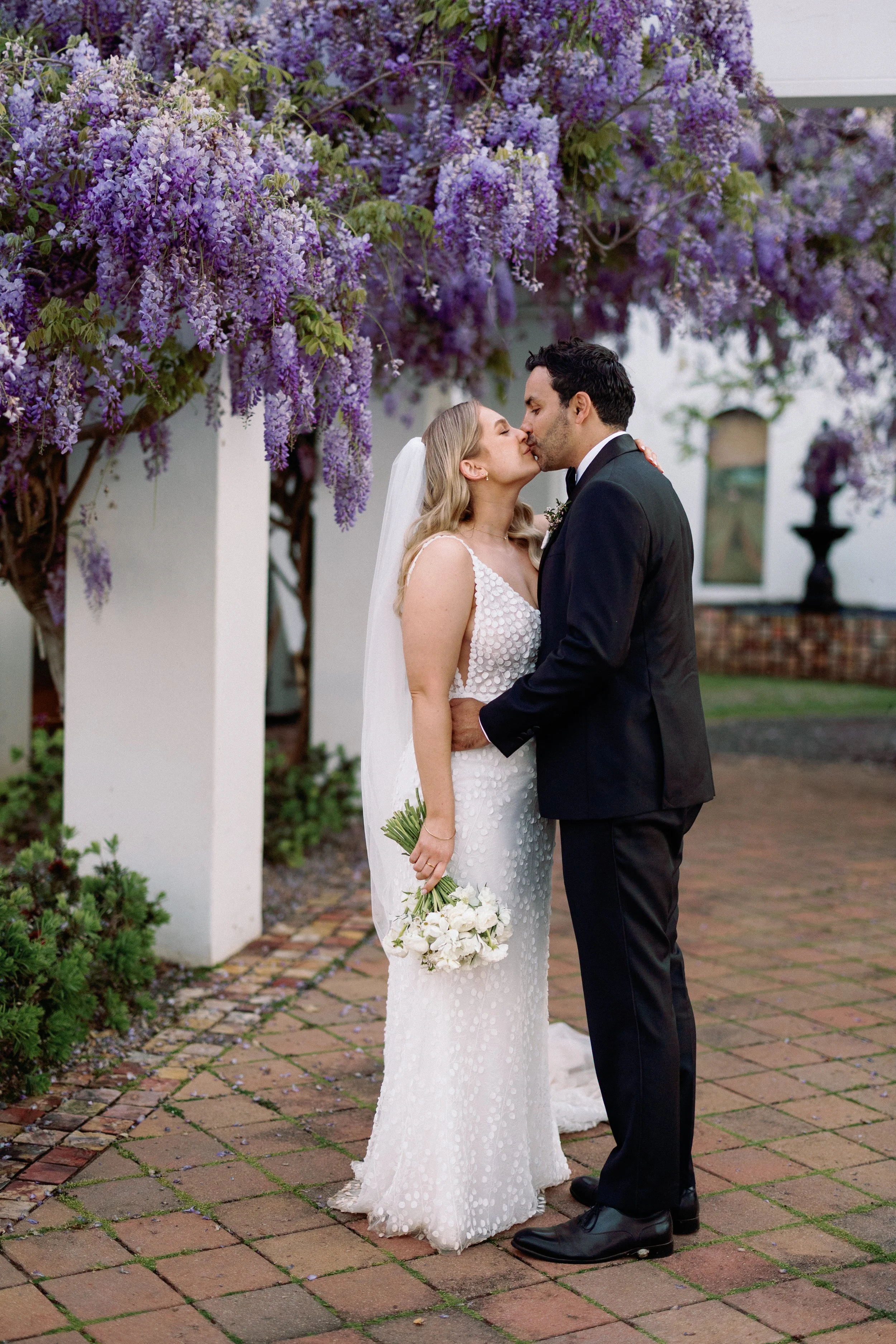 Bride and groom sharing a kiss under purple flowering wisteria at their wedding.