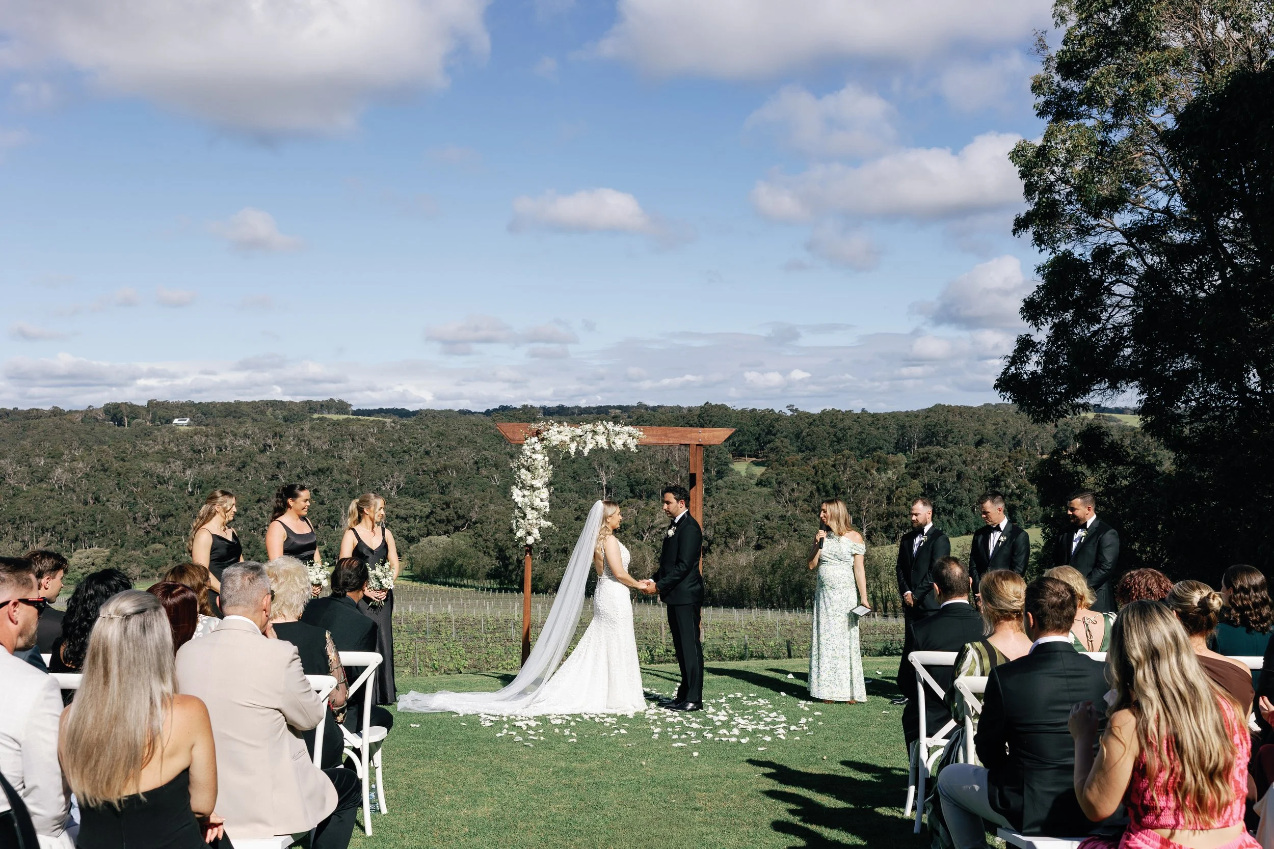 A wedding ceremony outdoors with the bride and groom holding hands, surrounded by bridesmaids, groomsmen, and guests, with a scenic rural landscape in the background.