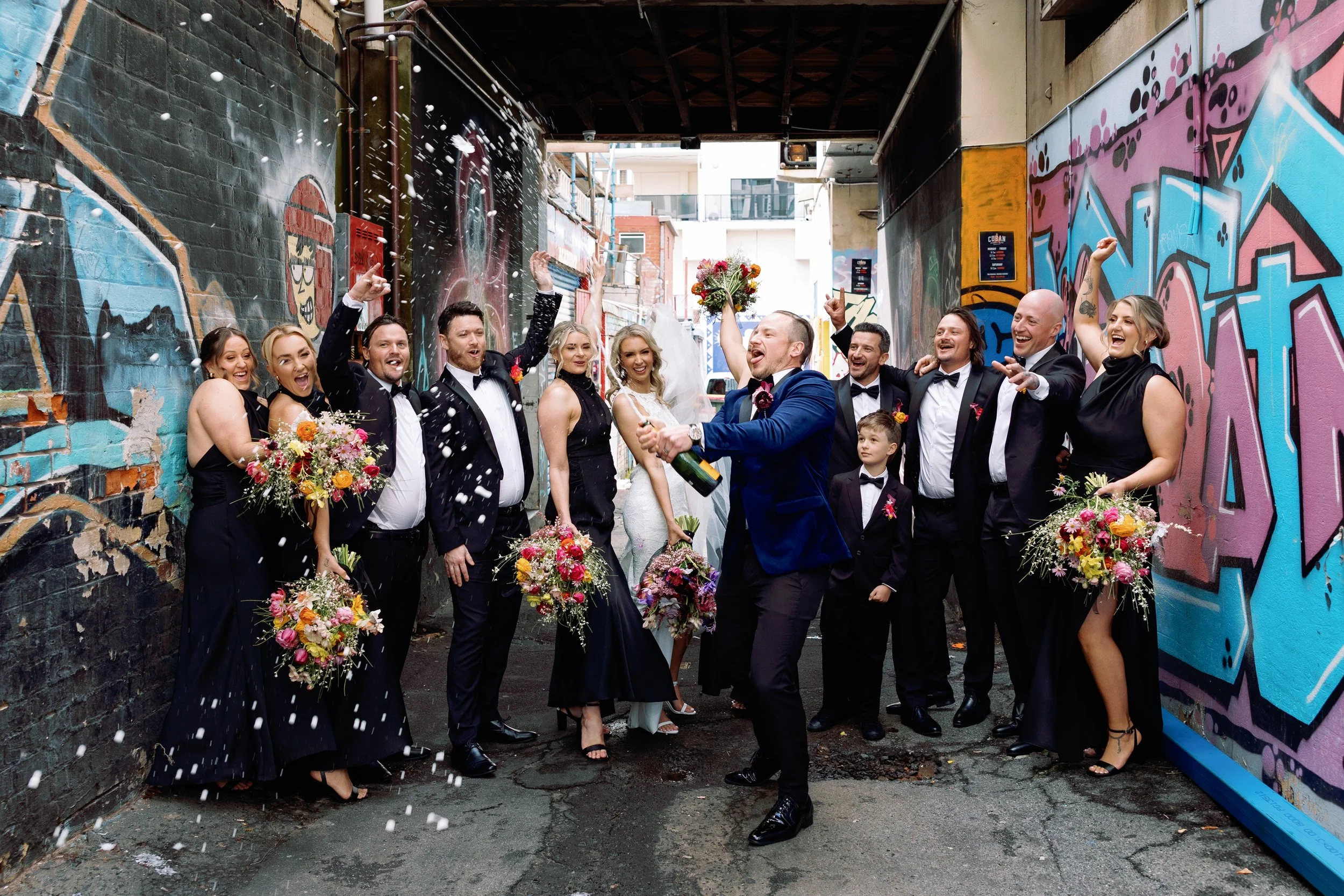 Group of people in formal attire celebrating, with some holding bouquets of flowers, in an alleyway with graffiti walls, as a man sprays champagne.