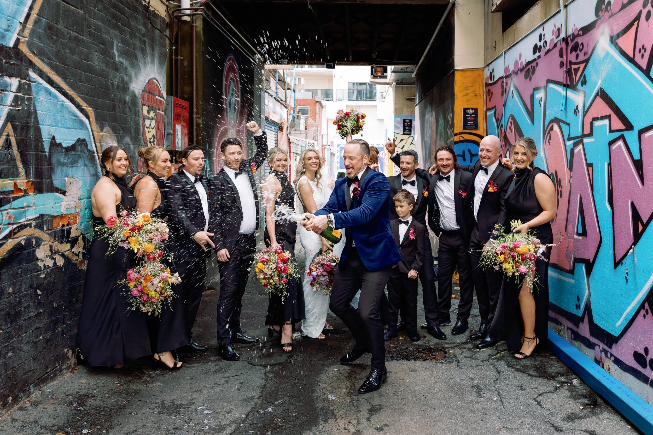 A group of wedding party members celebrating in an alley with colorful graffiti, with a man in a blue suit spraying champagne while others smile and hold bouquets.