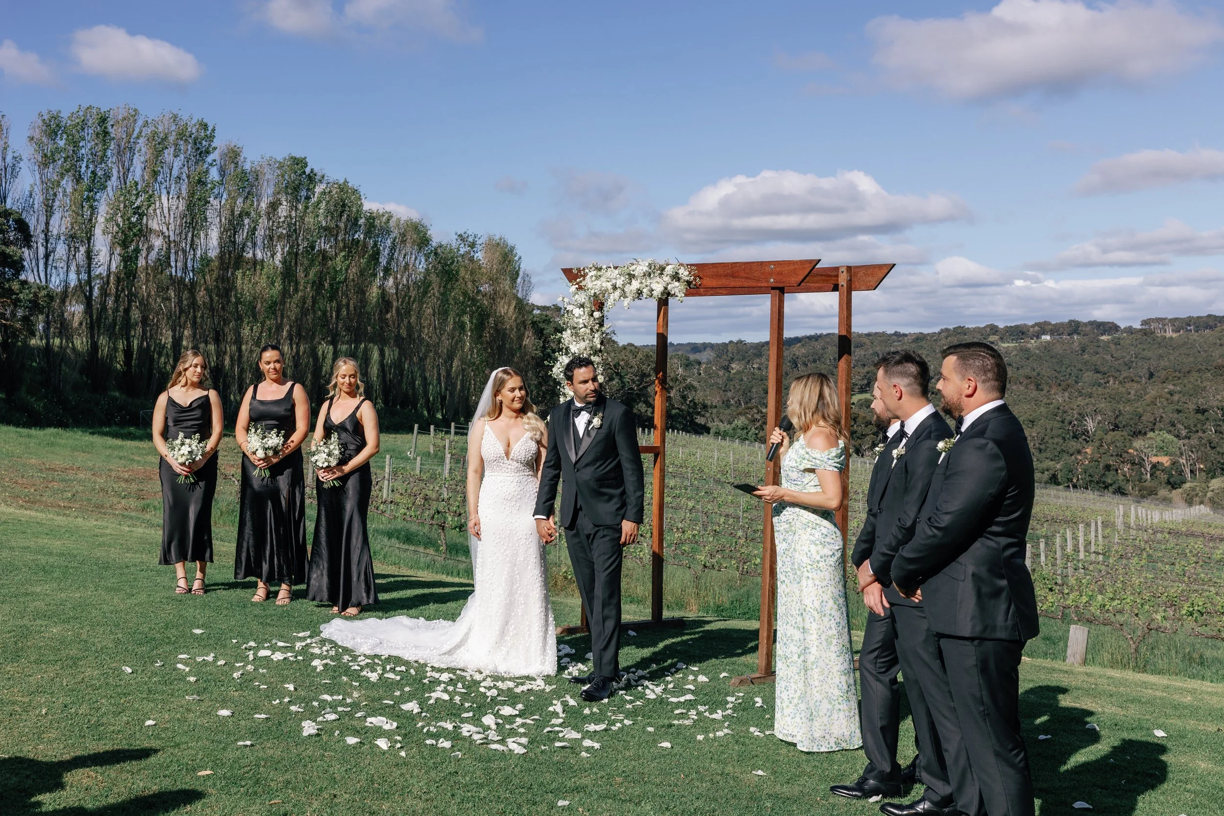 A wedding ceremony taking place outdoors with a bride and groom holding hands, surrounded by bridesmaids, groomsmen, and a officiant reading from a microphone under a decorative wooden arch, with a vineyard and trees in the background on a sunny day.