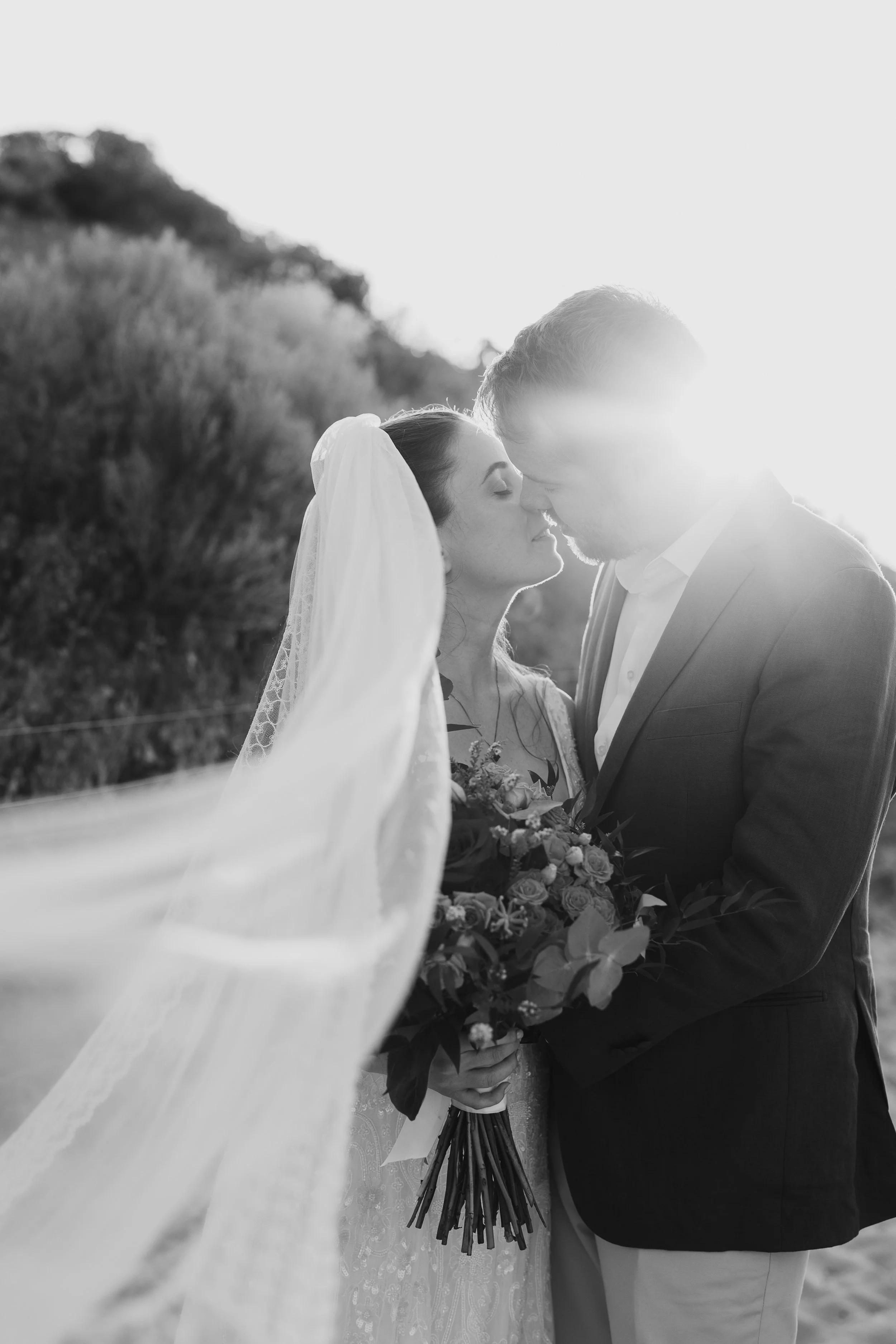 Black and white photo of a bride and groom with foreheads touching, the bride holding a bouquet, backlit by the sun, outdoors in a natural setting.