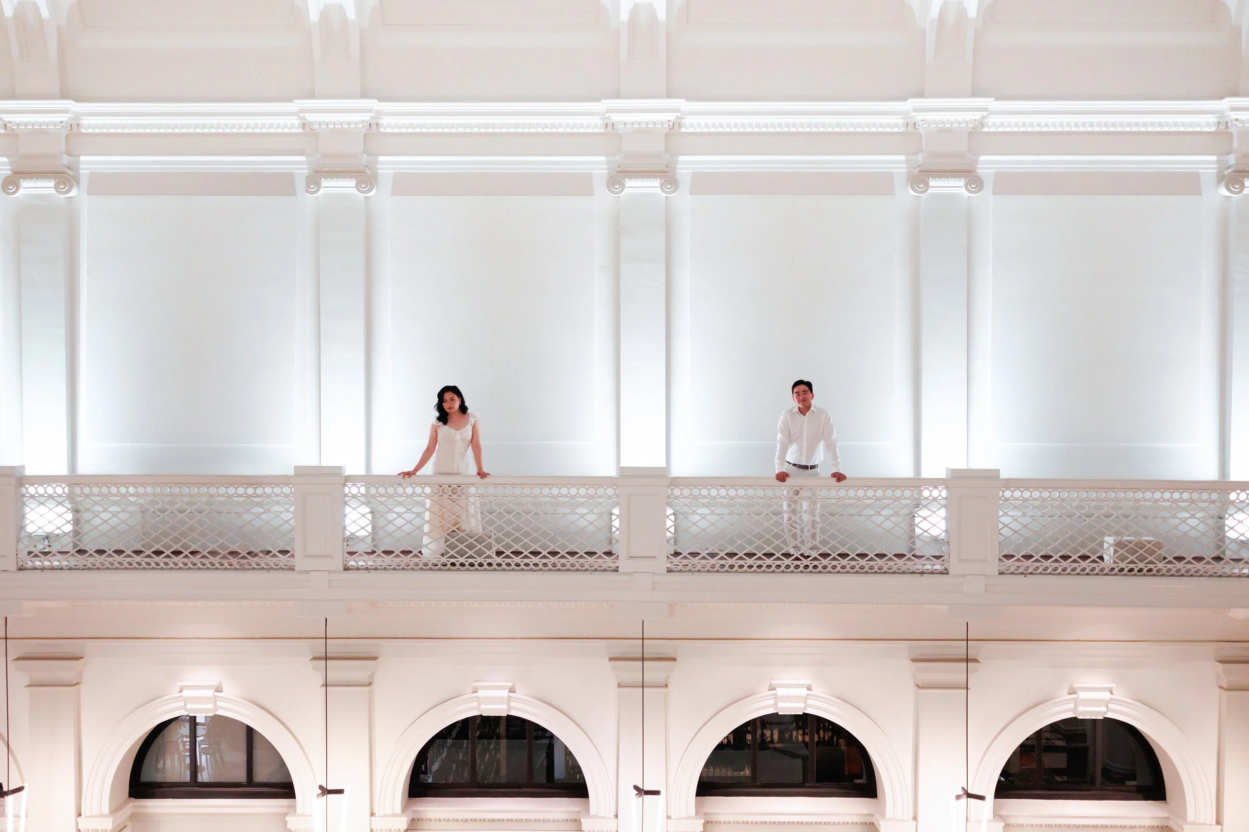 A woman and a man standing on a white balcony inside a bright, elegant building with tall windows and detailed architectural design.
