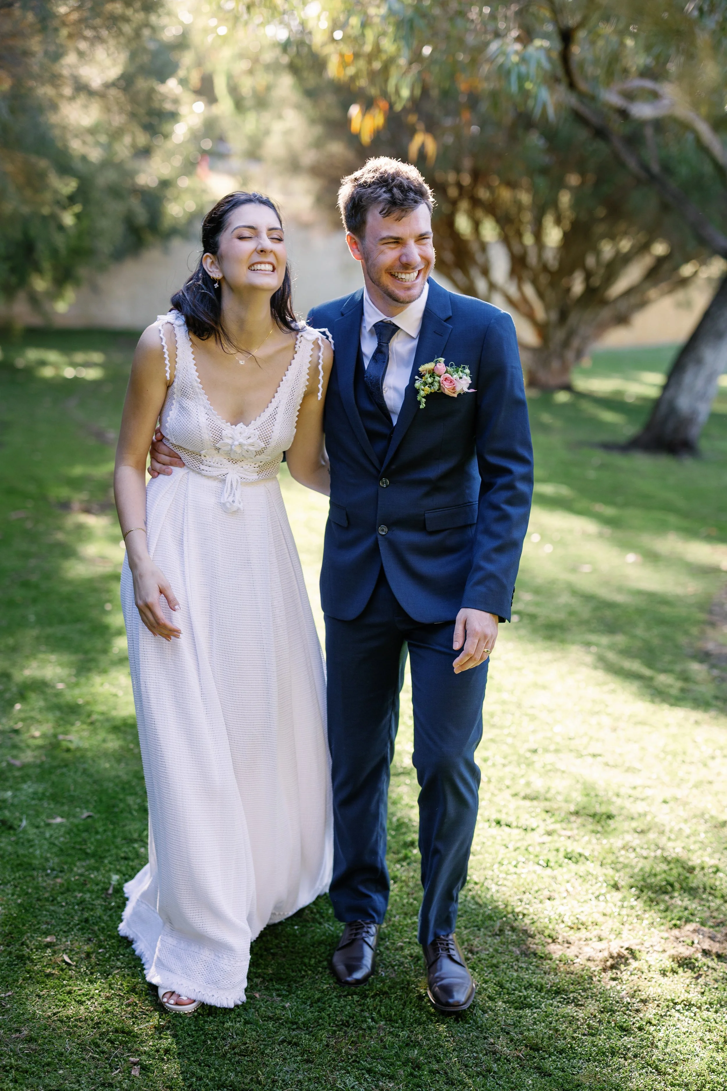 A smiling bride and groom walk outdoors on a sunny day, surrounded by green trees and grass.