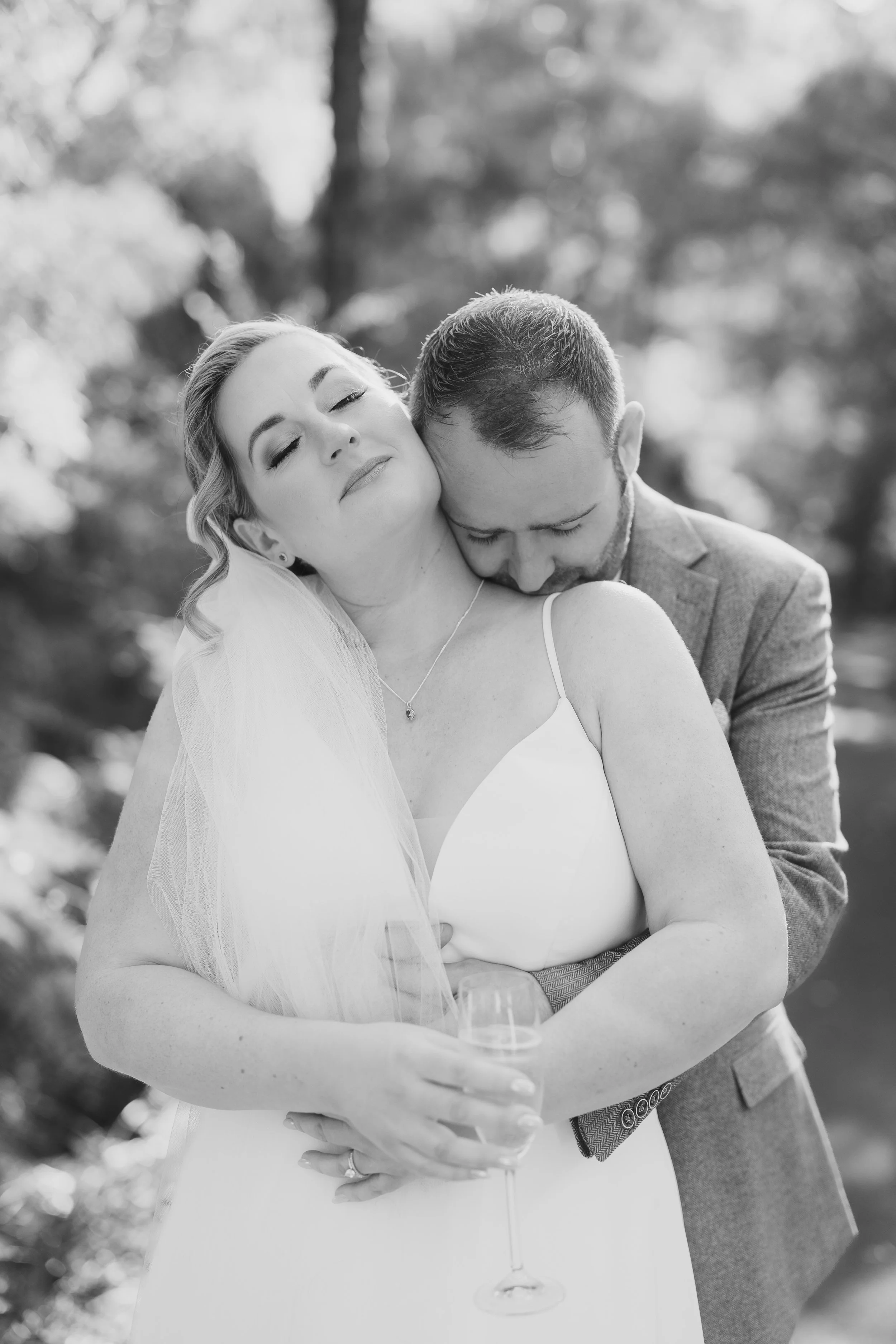 A black and white photo of a bride and groom embracing outdoors, the bride holding a glass of champagne with her eyes closed and the groom resting his head on her shoulder.