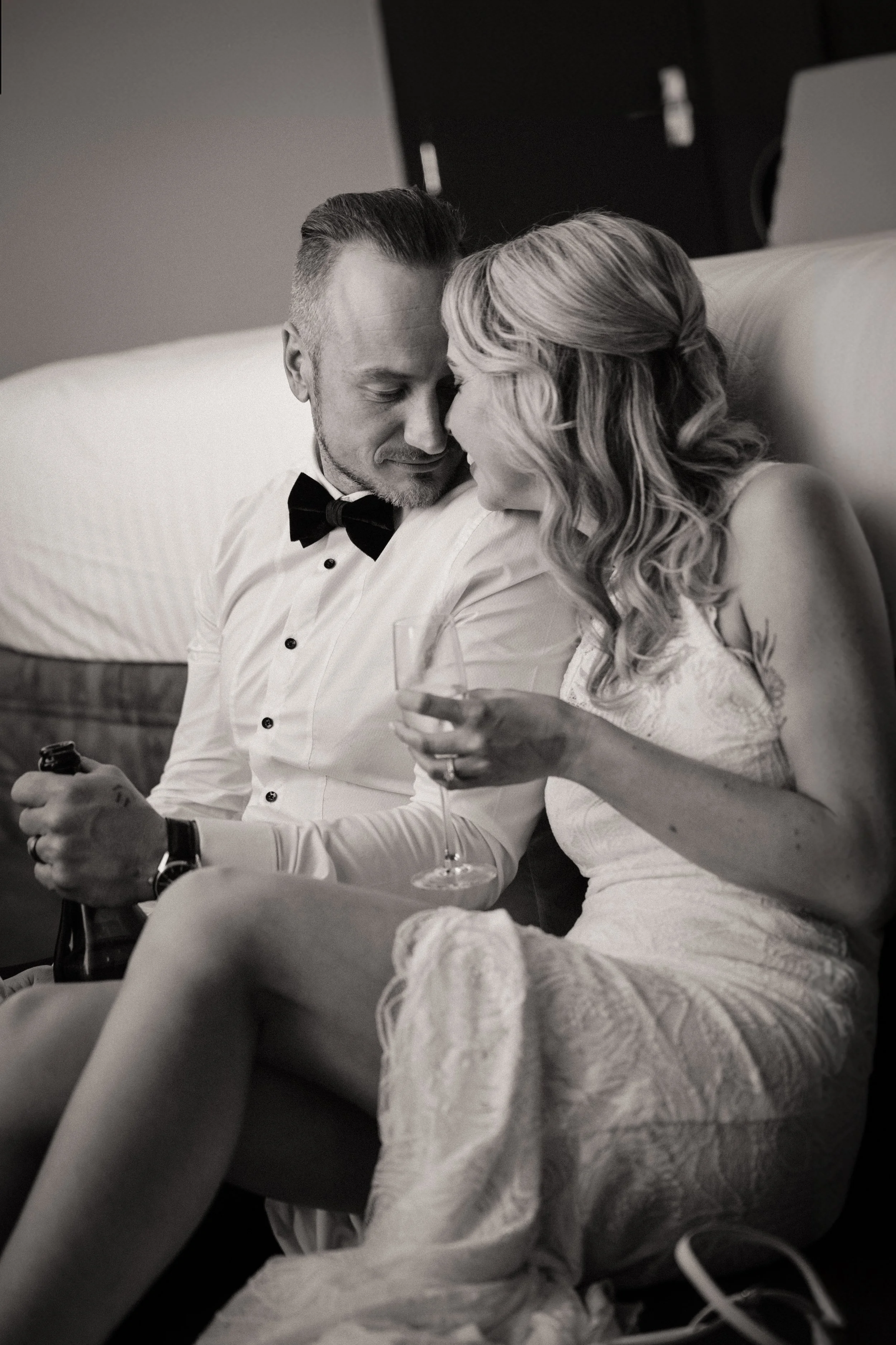A black-and-white photo of a couple sitting closely on a bed, laughing and touching foreheads. The man is wearing a tuxedo with a bow tie, holding a bottle, and the woman is in a dress, holding a wine glass.