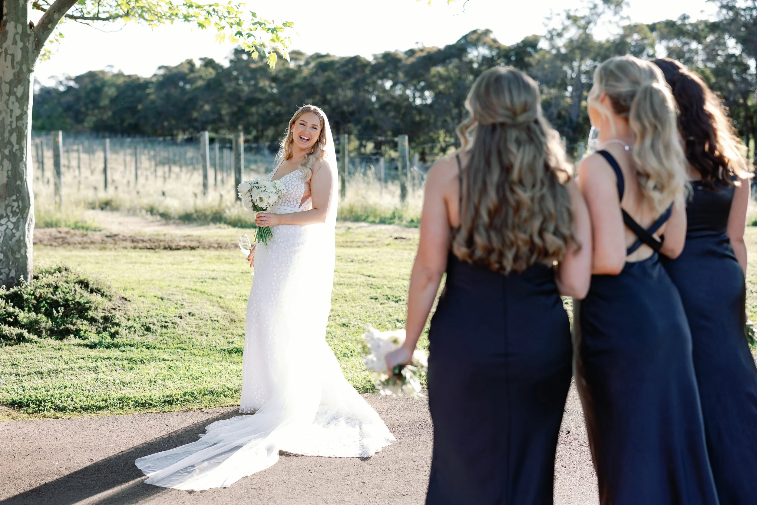 A bride in a white wedding gown holding a bouquet, smiling at her wedding ceremony outdoors, with three bridesmaids facing her, dressed in dark dresses.