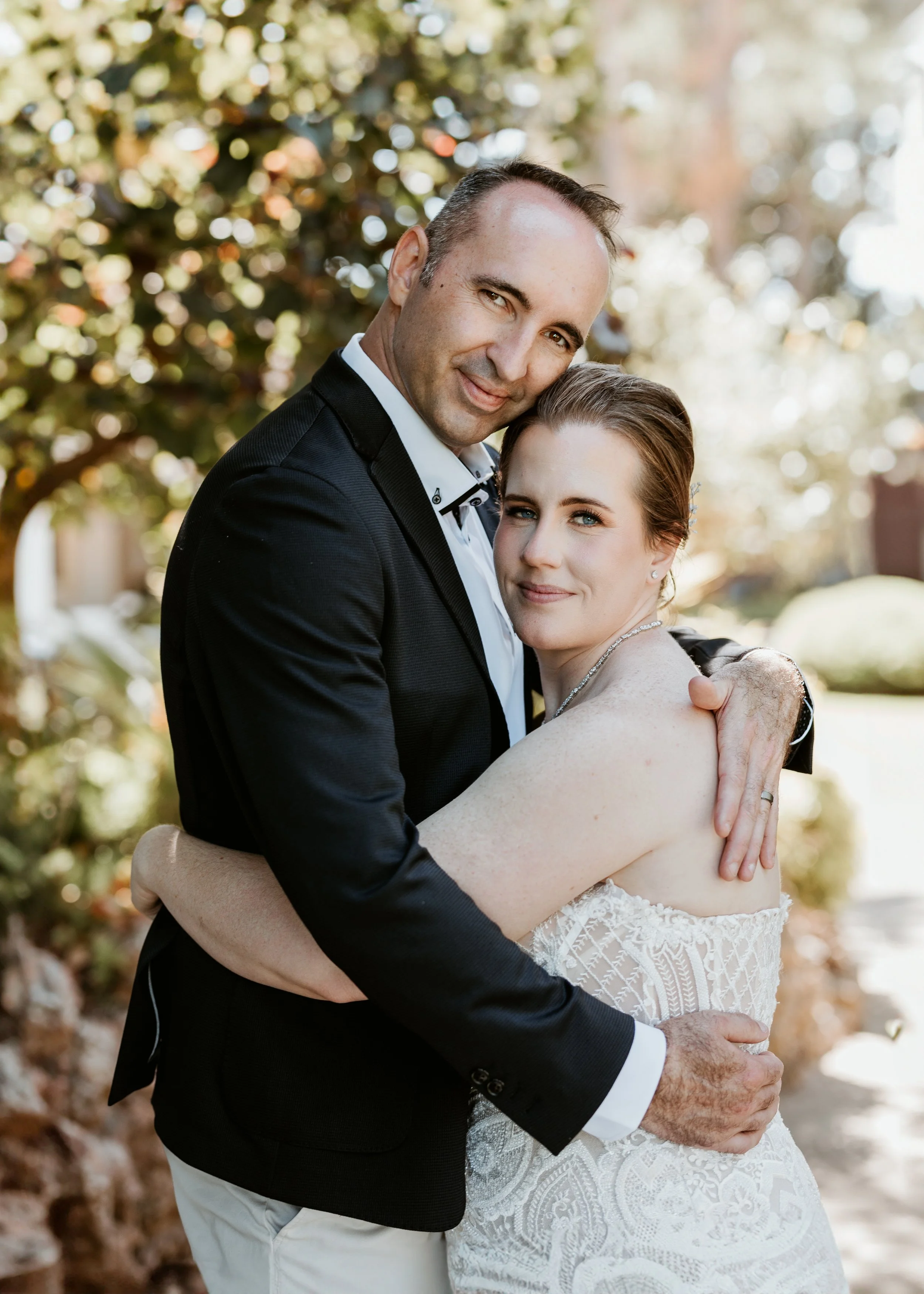 A couple in wedding attire embracing outdoors with trees and bokeh lights in the background.