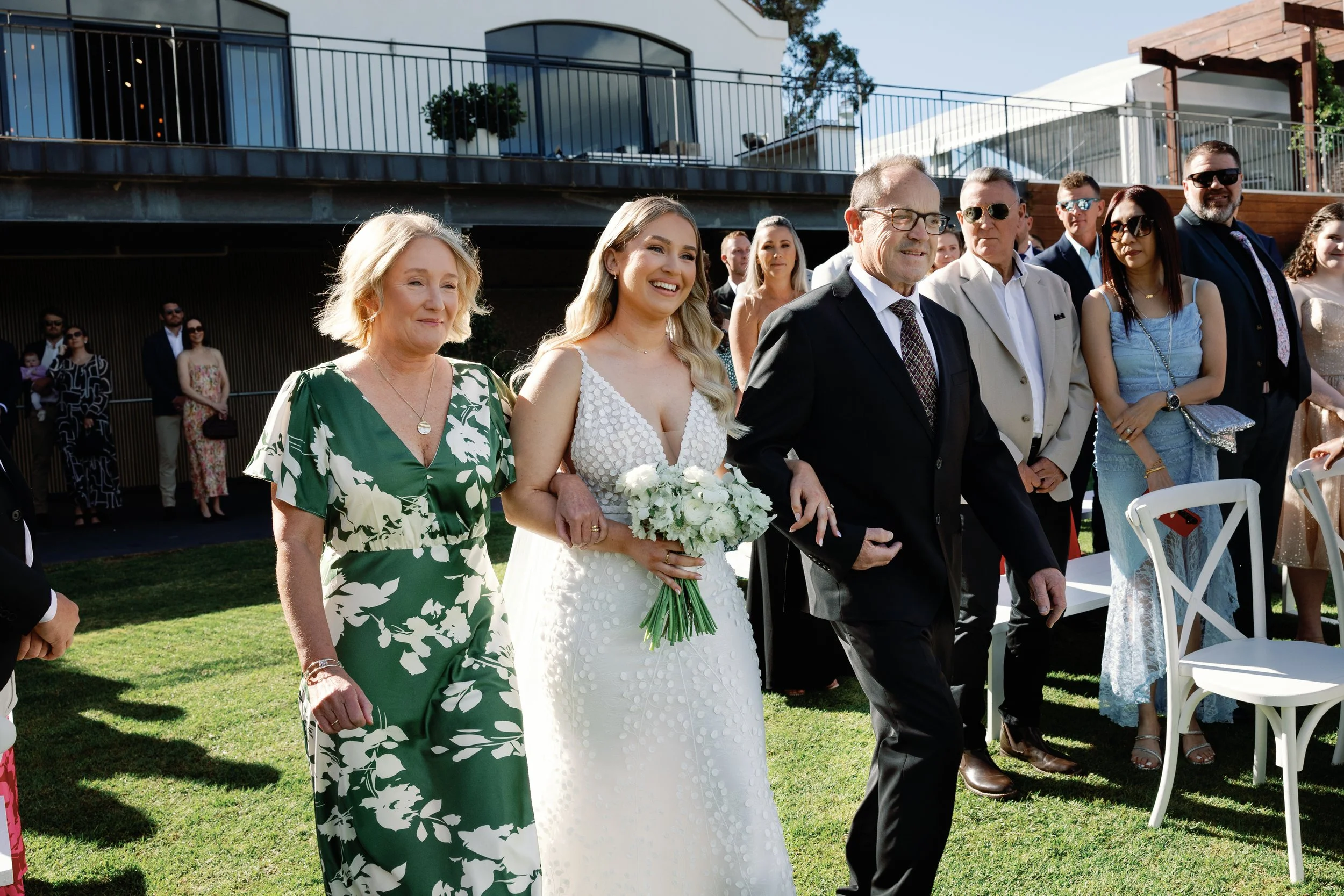 A bride in a white dress holding a bouquet of white flowers is walking with her father, who is dressed in a black suit, during an outdoor wedding ceremony. There are multiple guests standing and watching in the background.
