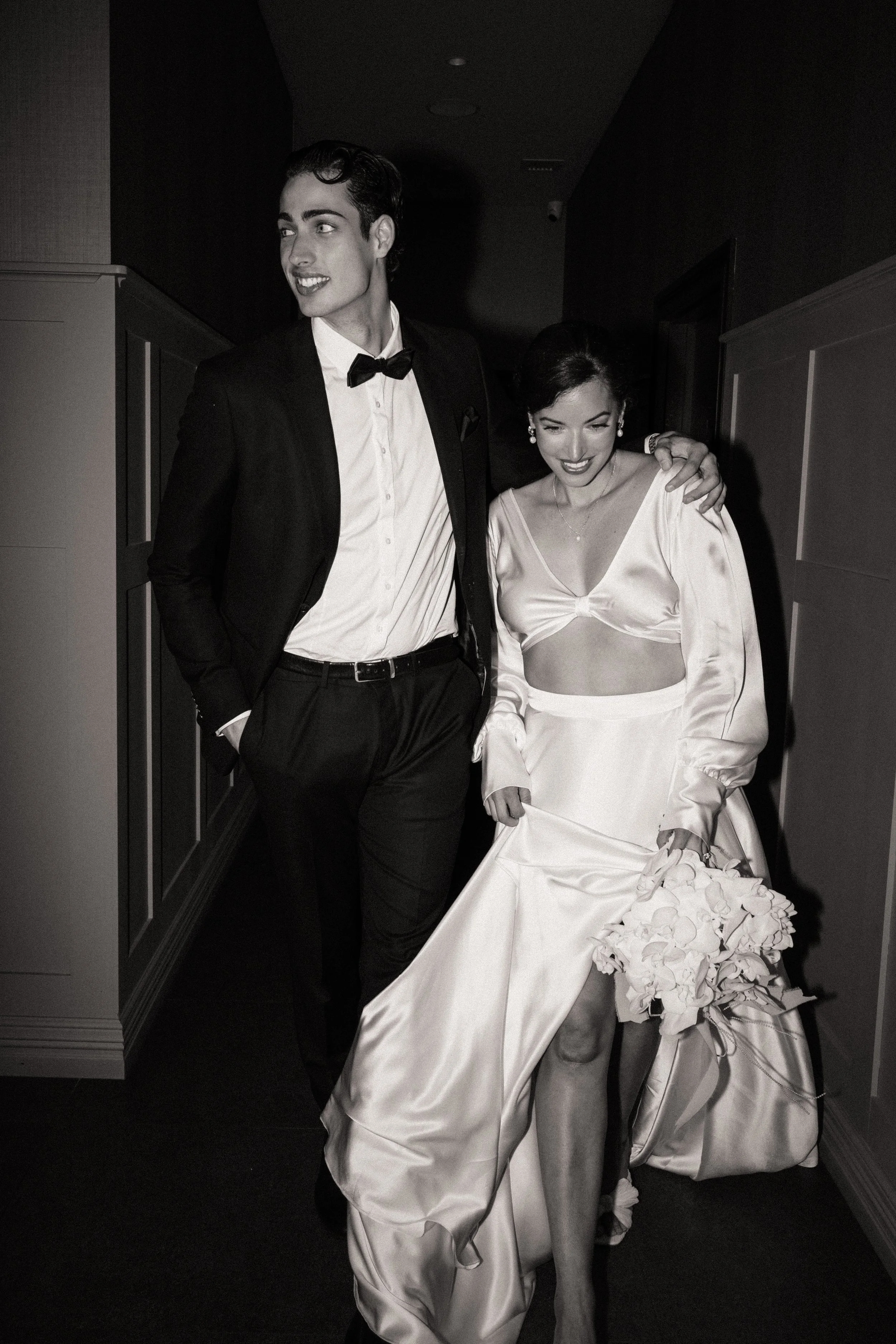A black-and-white photo of a man and woman walking together, the woman holding a bouquet of flowers, dressed in formal attire, smiling and enjoying the moment.