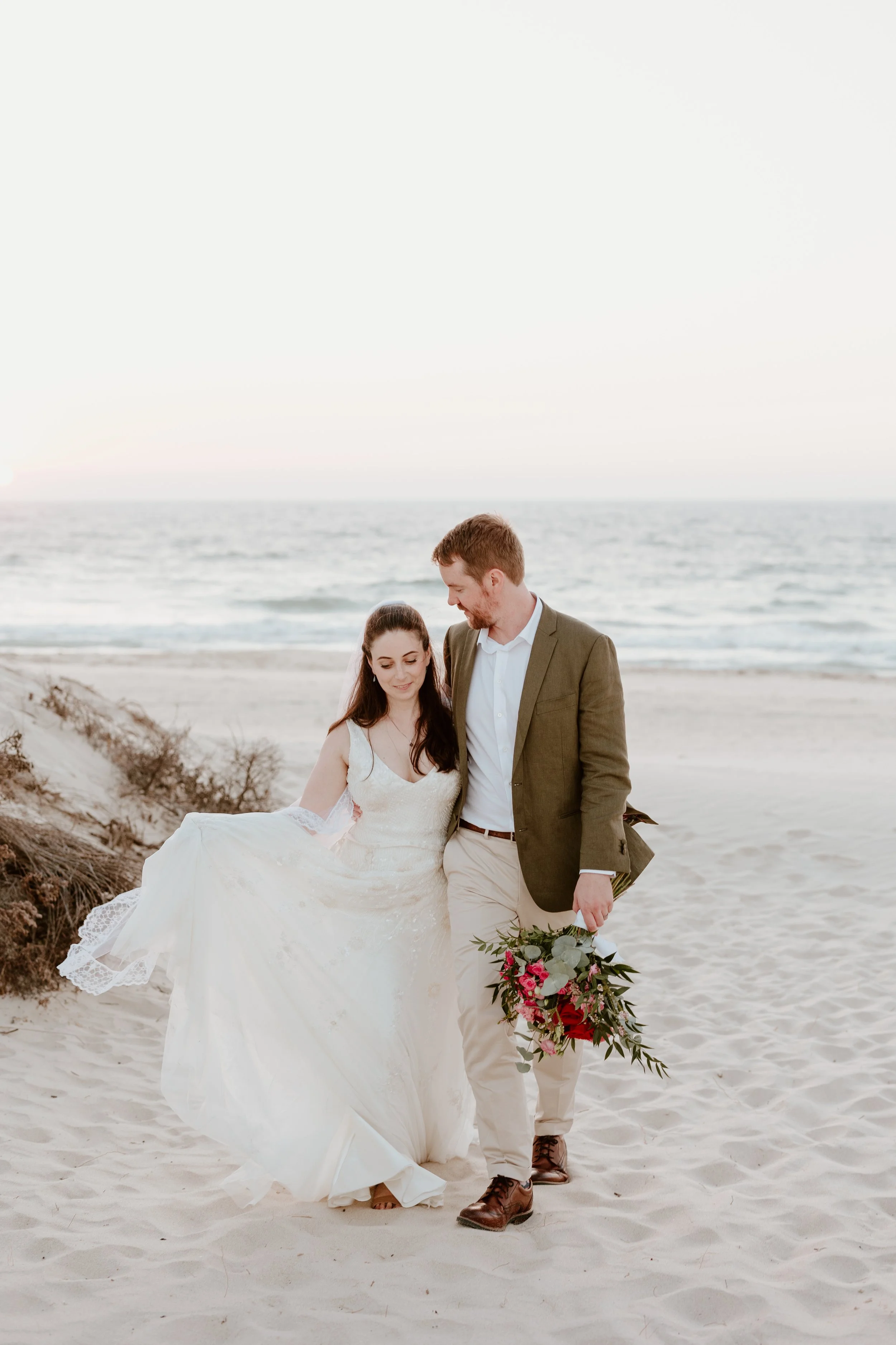 A bride in a white wedding gown and a groom in a beige suit walk on the beach, holding a bouquet, during sunset.