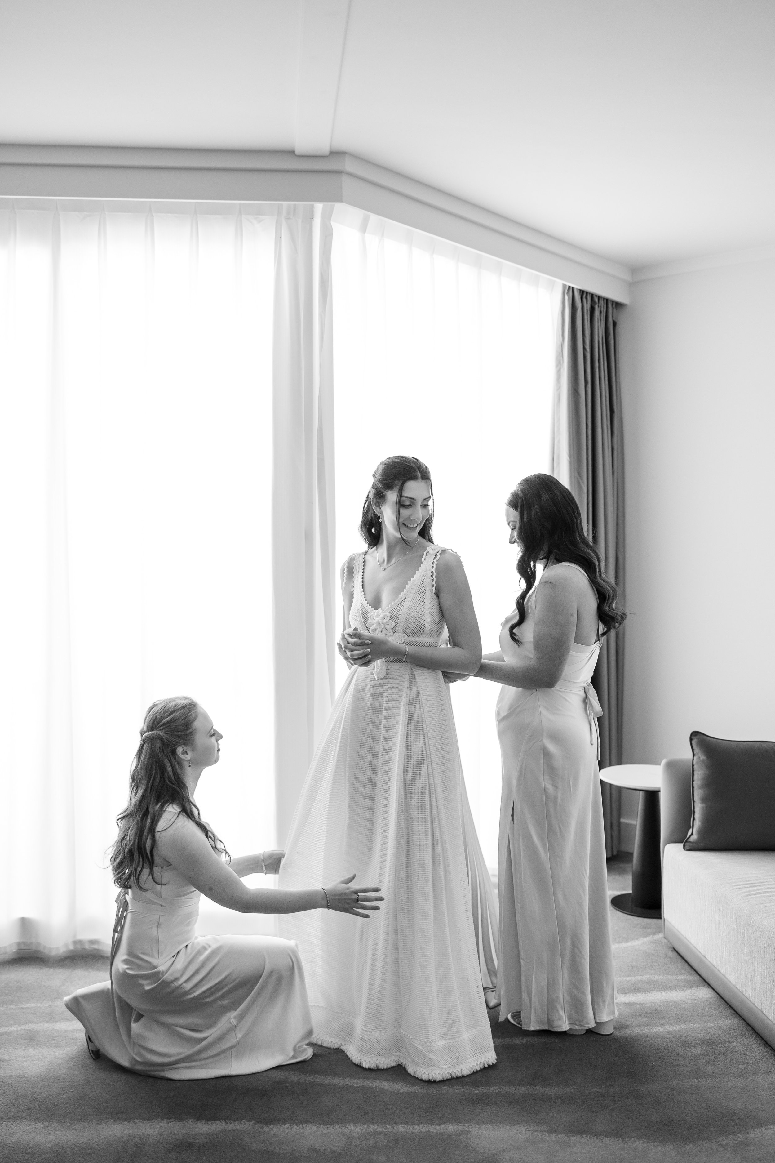 Black and white photo of three women in a hotel room, two standing and one kneeling, preparing a bride for her wedding, with a bright window in the background.