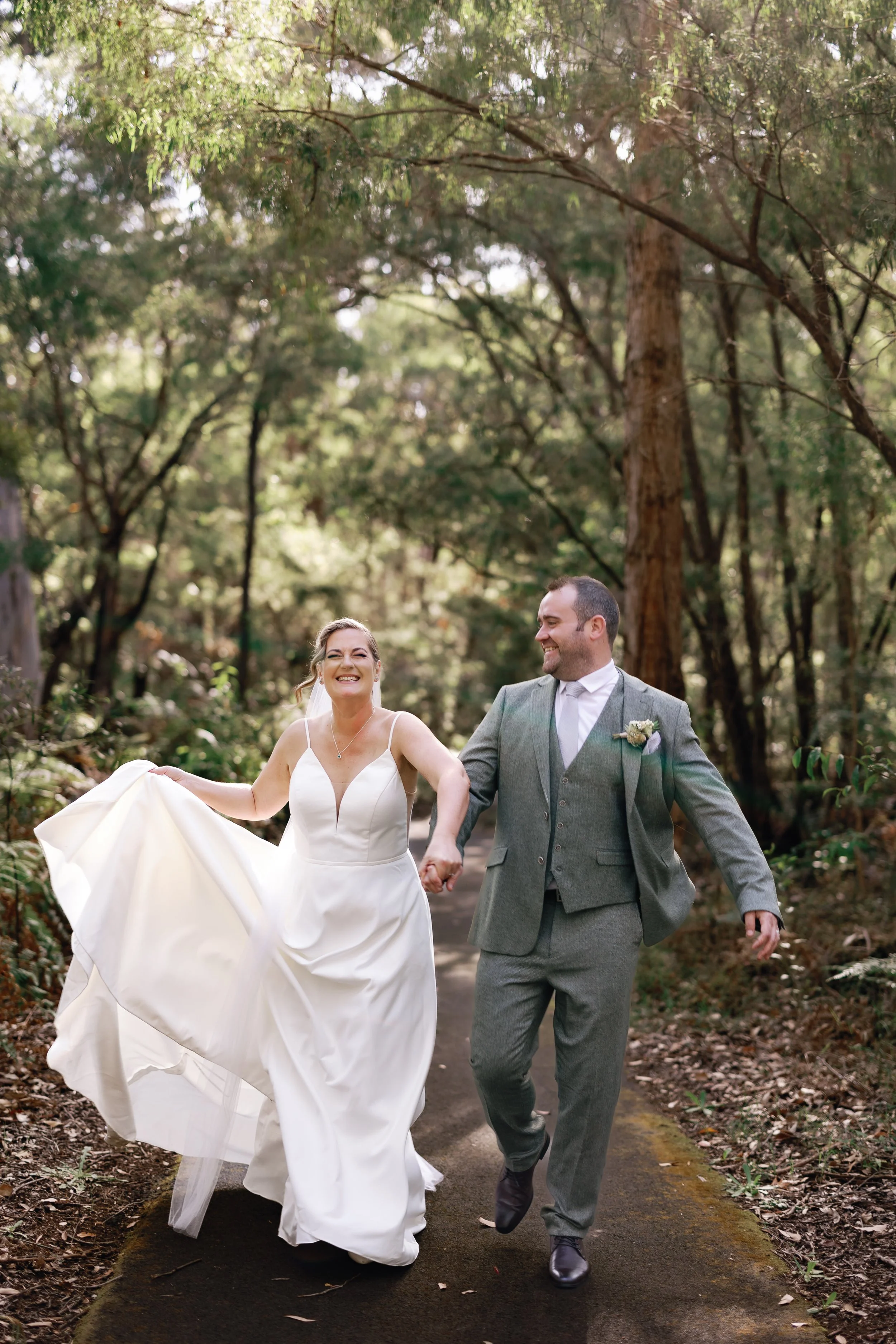 A smiling bride and groom holding hands and walking through a wooded path during their wedding day.