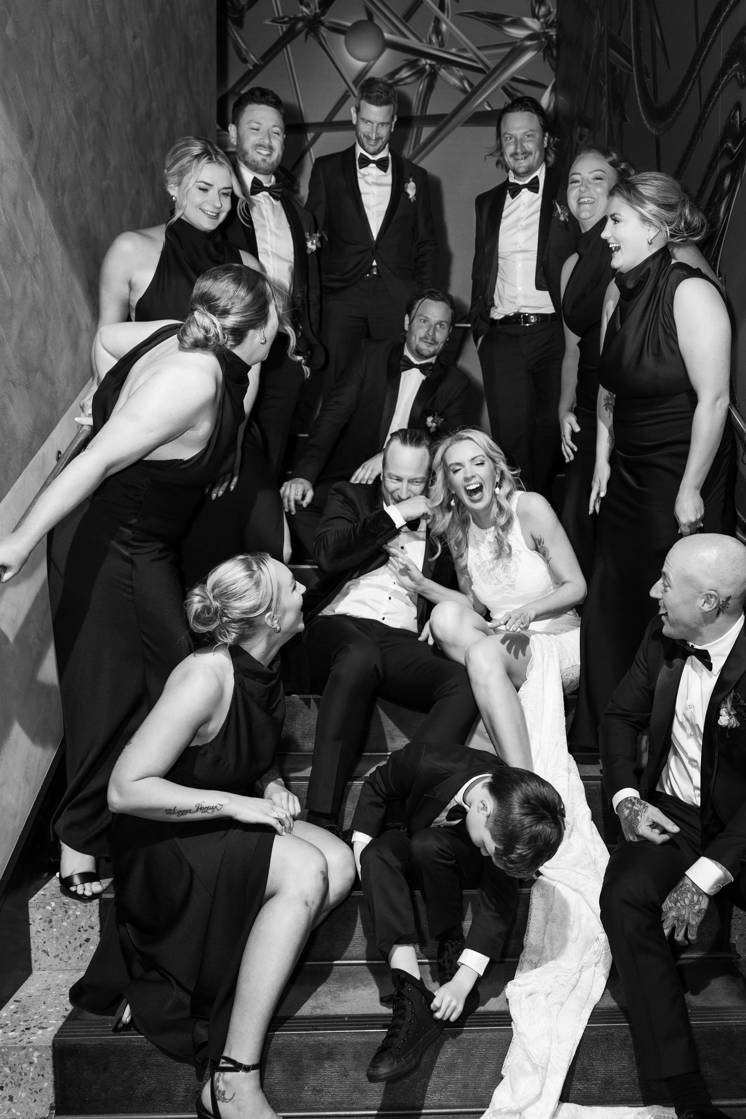 A group of people dressed in formal attire, gathered on a staircase, celebrating and laughing at a wedding or special event, with some sitting and others standing around at QT Hotel Perth 