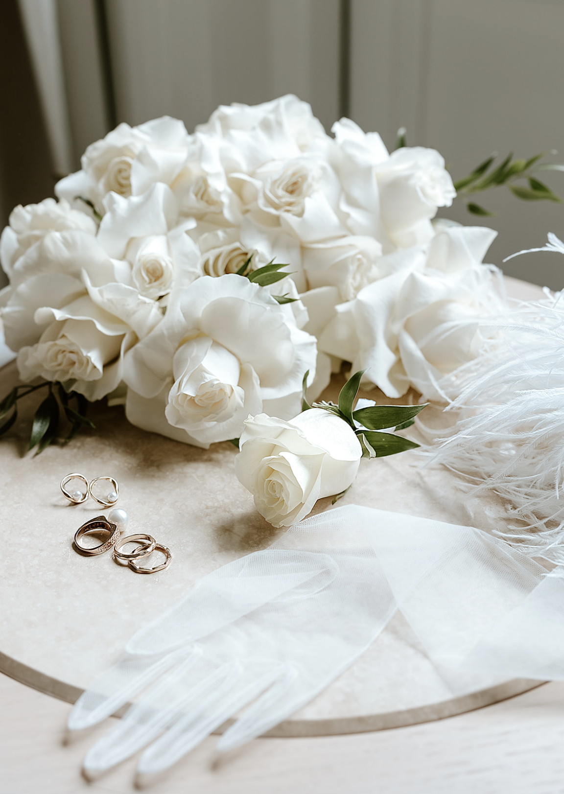 Courtly collection of white roses, calla lilies, and greenery, with jewelry and white gloves on a marble surface.