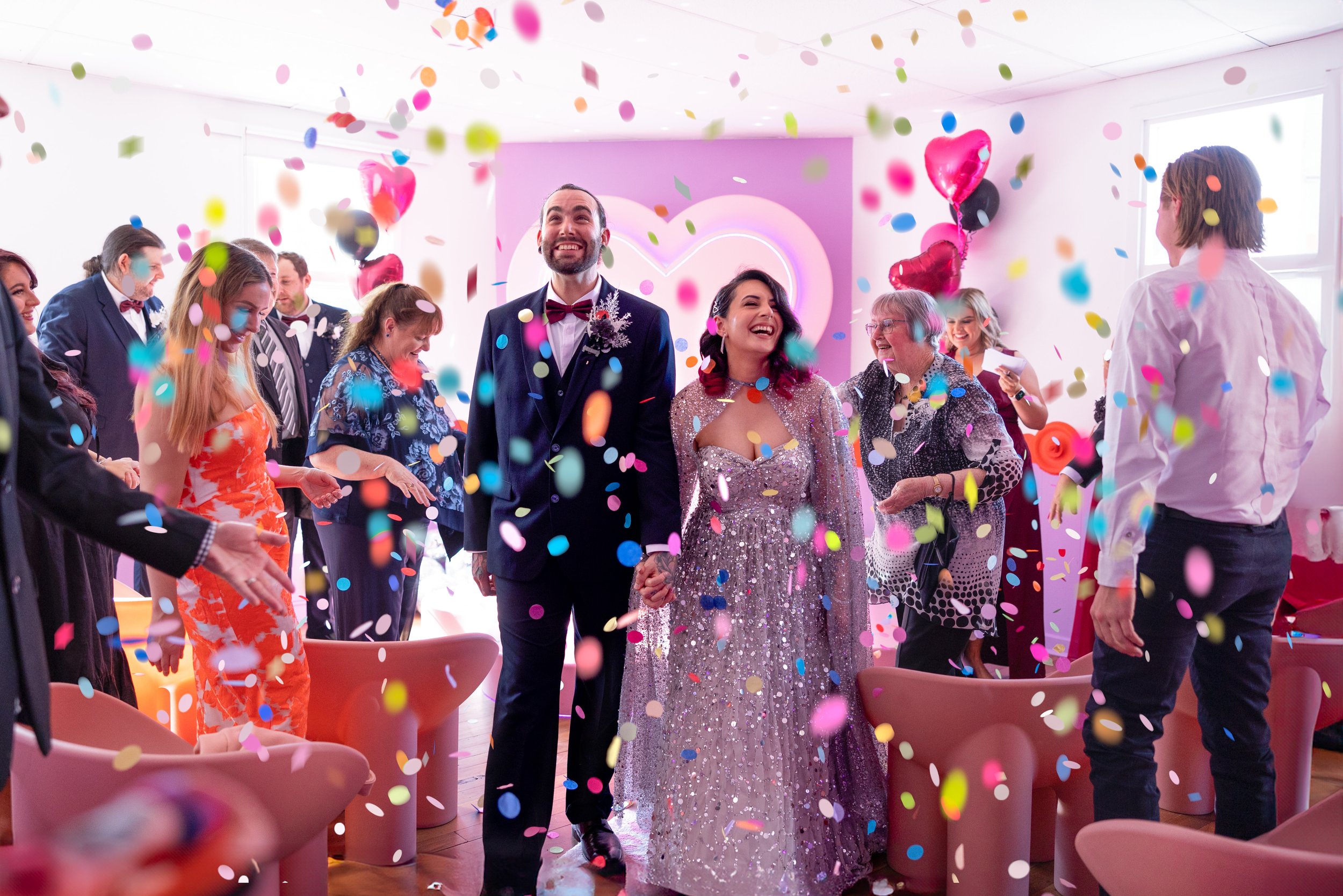A wedding celebration with a bride and groom holding hands and smiling as they walk into the room, surrounded by guests and colorful confetti. The bride wears a sparkly gown, and the groom wears a suit. The room is decorated with pink heart-shaped ba