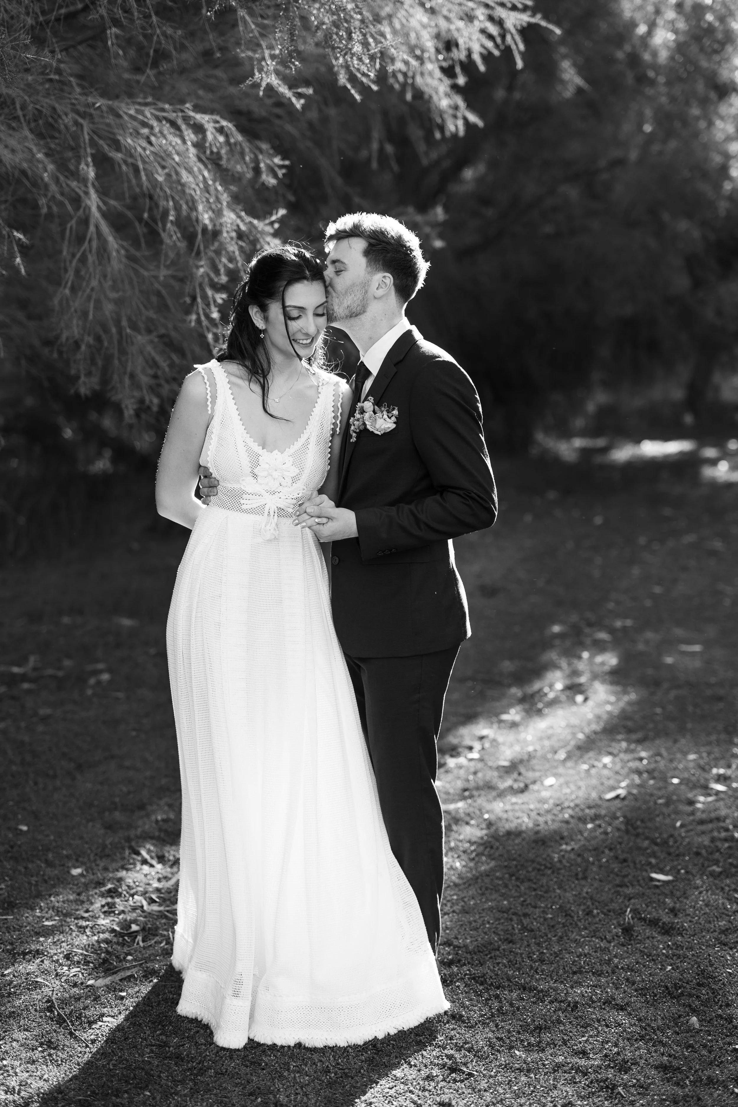 Black and white photo of a newlywed couple standing outdoors, the groom gently kissing the bride's forehead. The bride is wearing a white dress, and the groom is in a black suit with a boutonniere. Trees and sunlight are in the background.