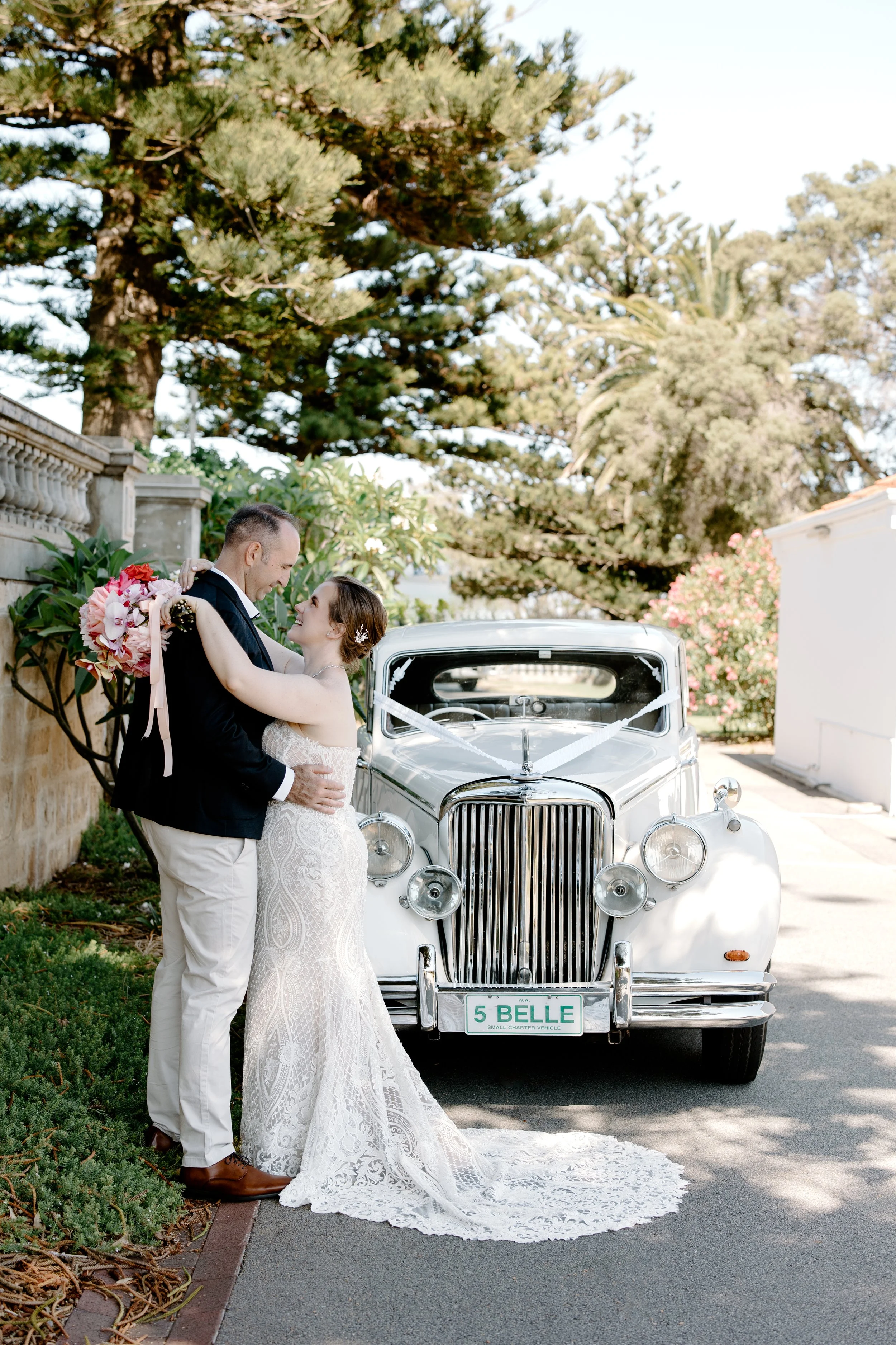 A bride and groom sharing a moment in front of a vintage white car with a floral arrangement, outdoors under a large tree and pink flowering shrub.