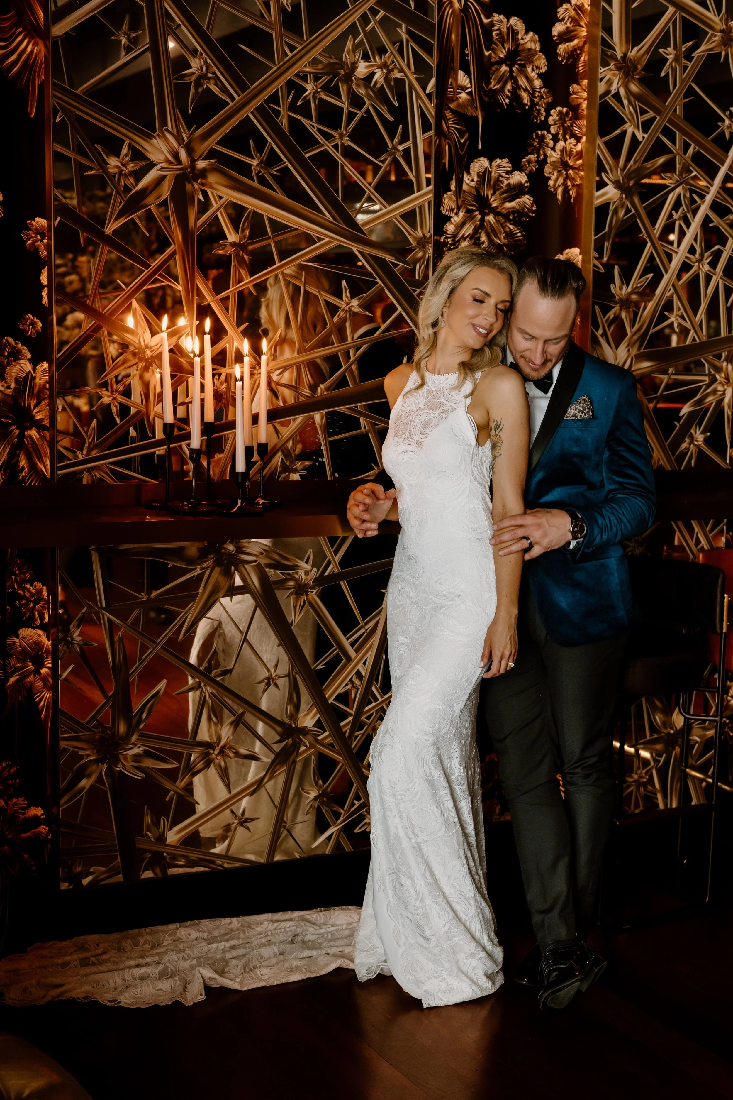 A newlywed couple embraces in an elegant, modern setting with a gold metallic geometric wall and a candle holder with lit candles in the background.